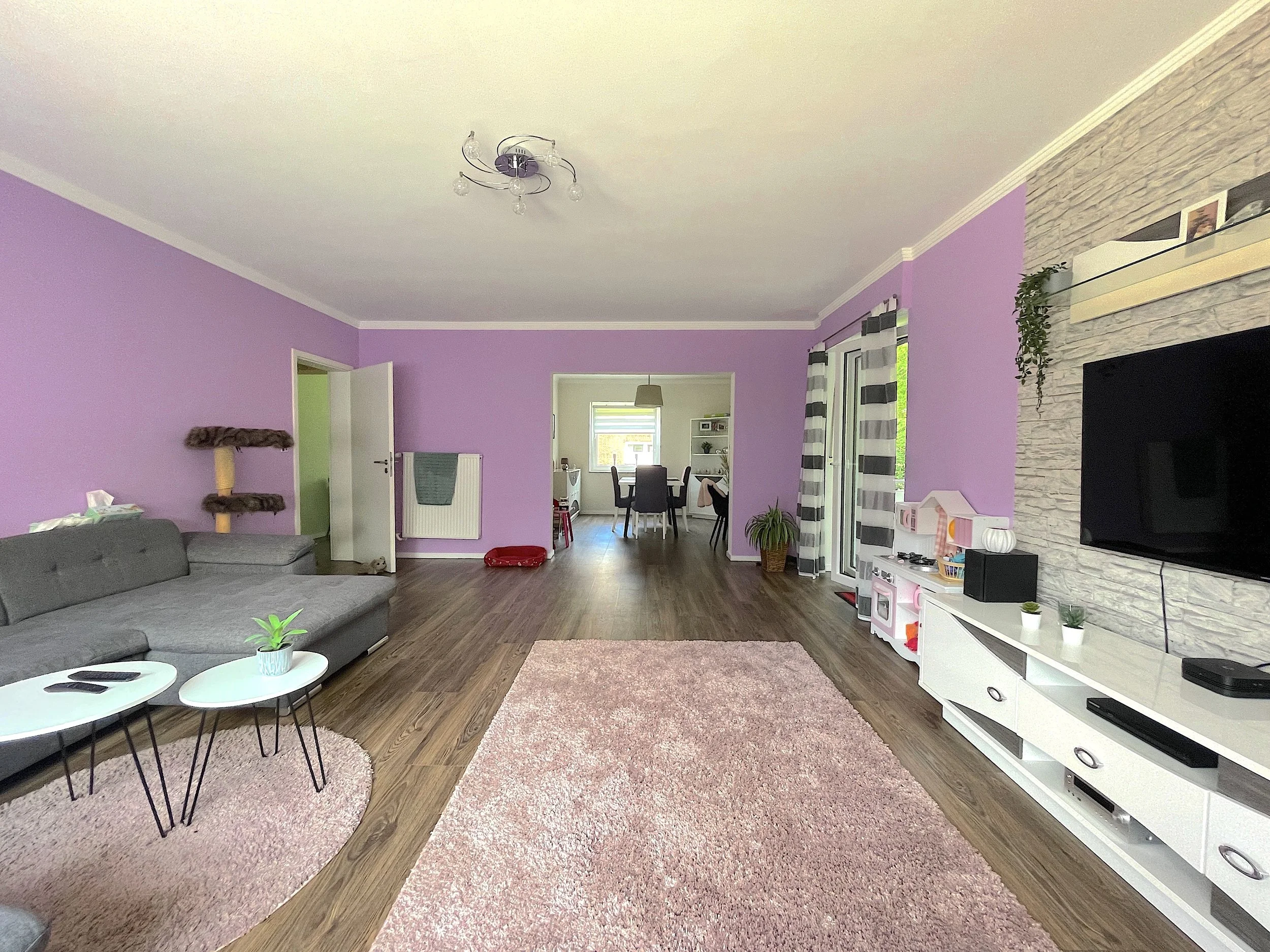 Gemütliches Wohnzimmer mit hellvioletten Wänden, grauem Sofa, weißen Couchtischen mit Pflanzen, Teppich, Fernseher an einer weißen Medienwand, im Hintergrund Essbereich mit Tisch und Stühlen, große Fenster mit weißen und schwarzen Gardinen, Holzfußboden.