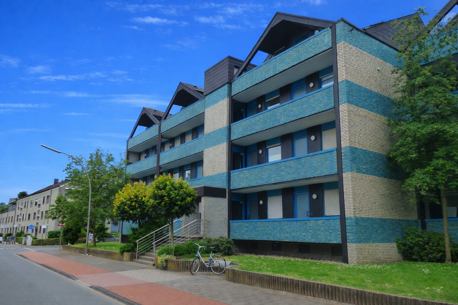 Modernes Mehrfamilienhaus mit grauem und blauen Fassaden, davor eine Straße, Bäume und ein Fahrrad.