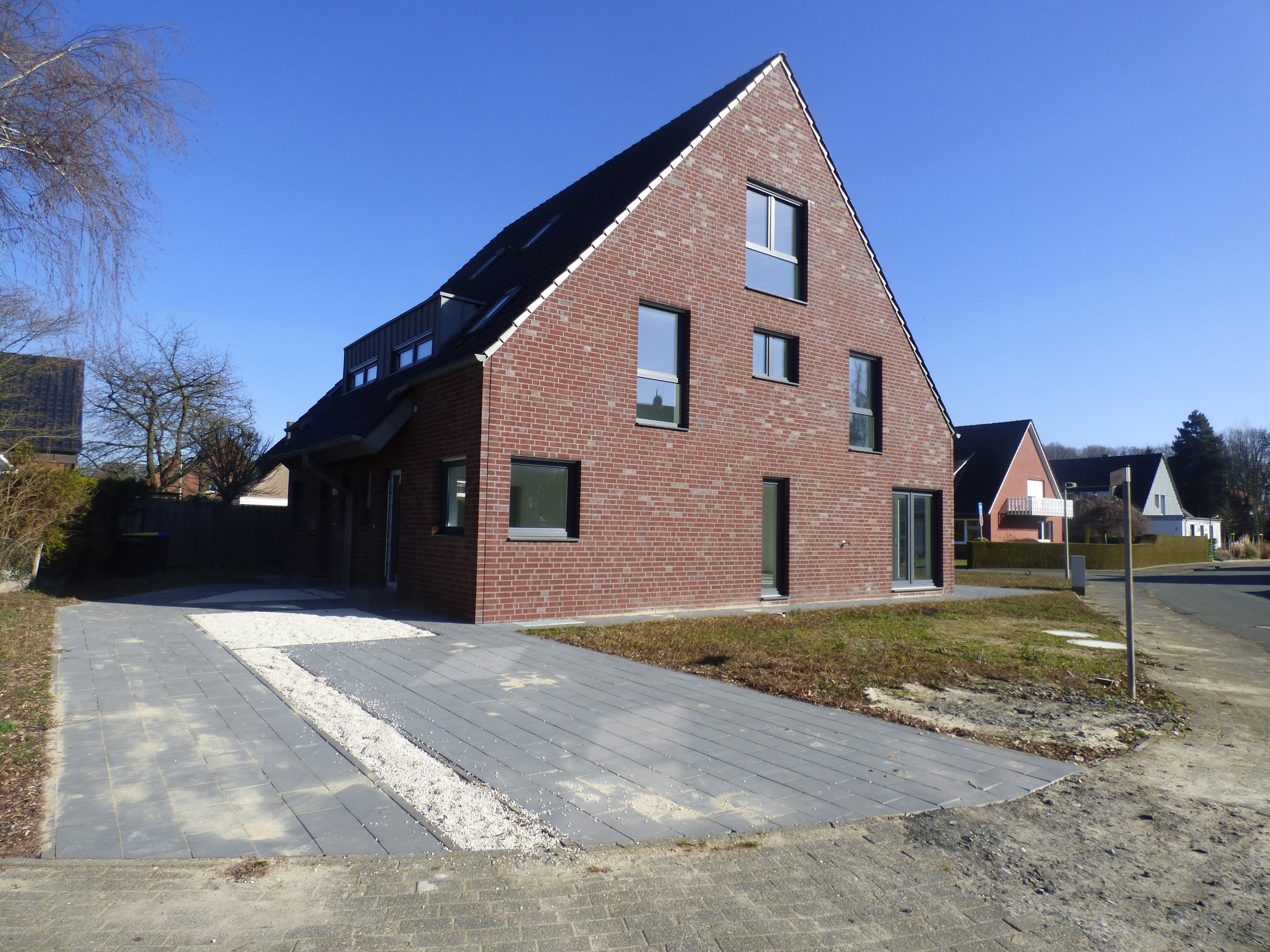 Modernes, rotes Backsteinhaus mit gewölbtem Dach und mehreren Fenstern in einer ruhigen Wohngegend bei sonnigem Himmel.