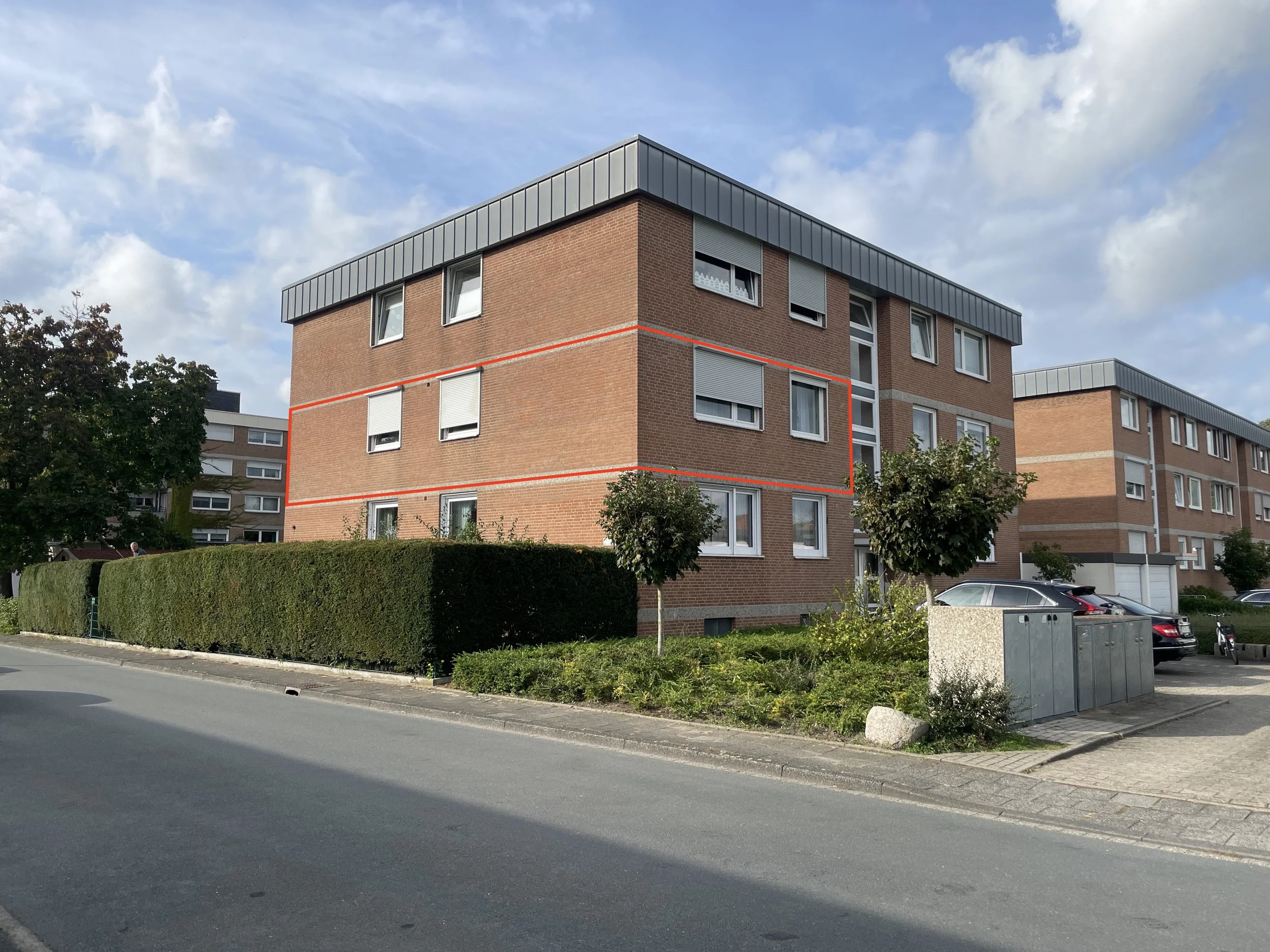Mehrfamilienhaus mit roten Ziegelfächen, Fenster und grauem Dach, umgeben von Bäumen, Hecken und parkenden Autos, bei sonnigem Himmel.