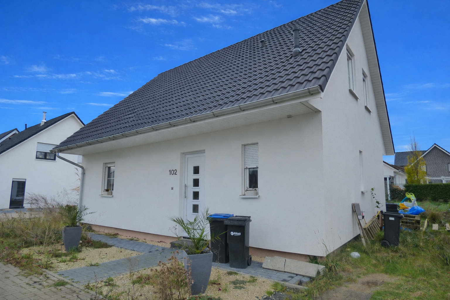 Ein weißes Einfamilienhaus mit grauem Satteldach und kleinen Fenstern, Nummer 102, mit grauem Gehweg und Garten mit Pflanzen und Mülltonnen im Vordergrund.