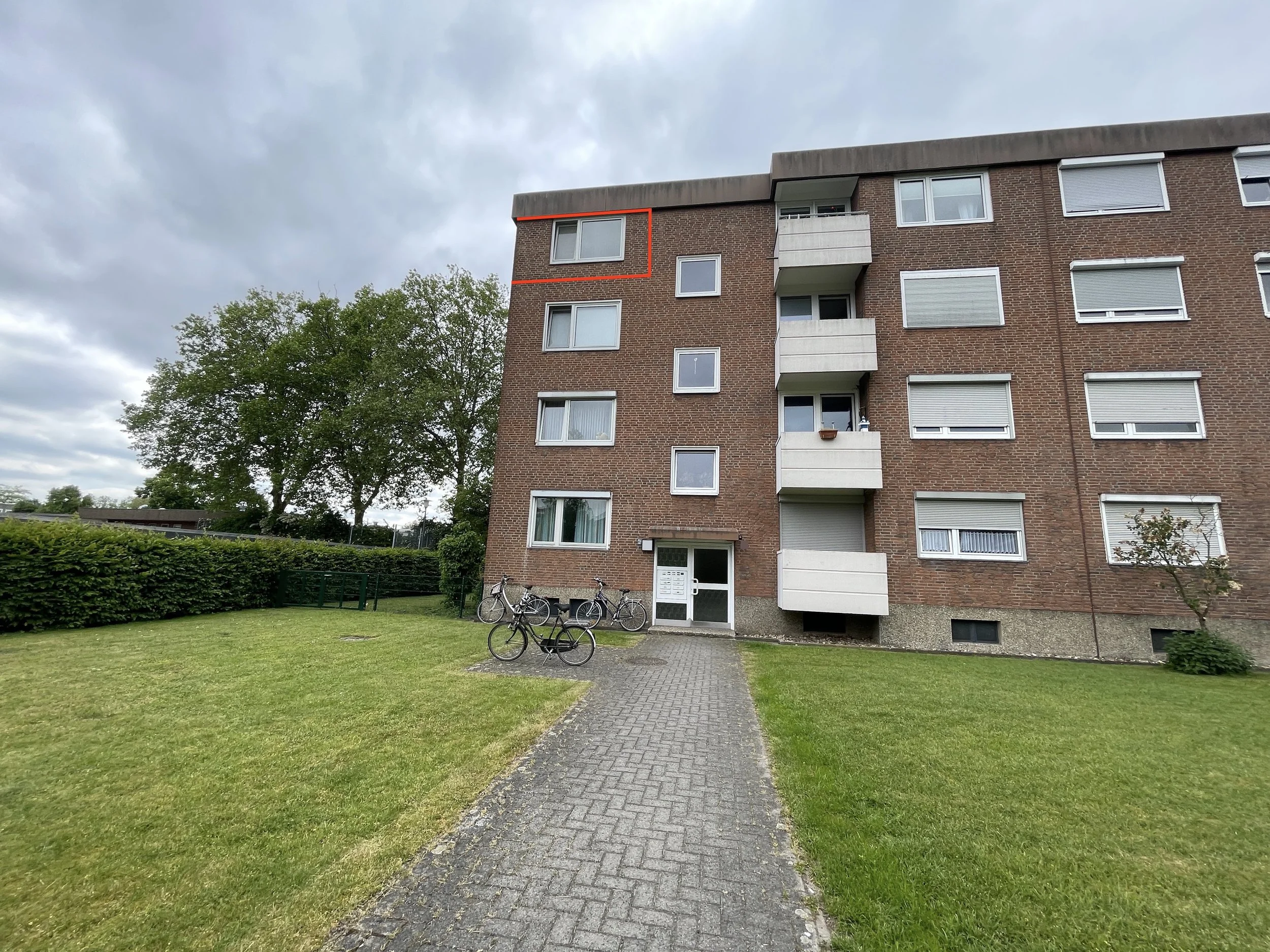 Mehrfamilienhaus mit vier Stockwerken, braune Ziegelmauer, weiße Balkone und Fenster, Fahrradständer mit Fahrrädern vor dem Eingang, grüner Rasen und Bäume in der Umgebung, bewölkter Himmel