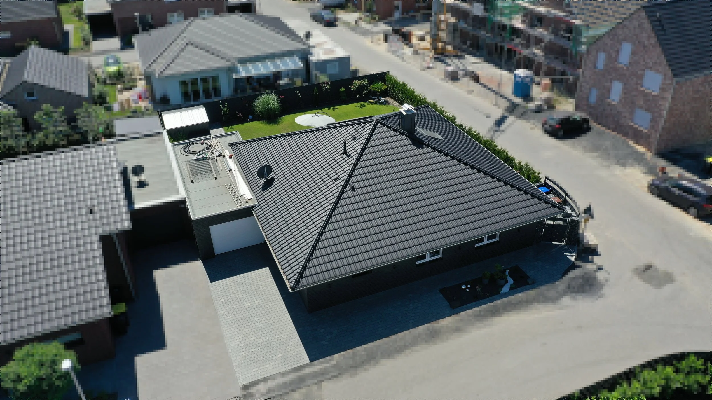 Luftaufnahme eines Hauses mit dunklem Dach, einer kleinen Gartenfläche mit Rasen, Bäumen und einem Sitzbereich, umgeben von Nachbarhäusern und Straßen.