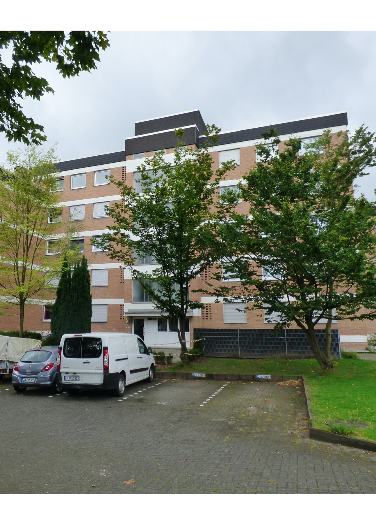 Mehrstöckiges Wohngebäude mit Bäumen im Vordergrund, Parkplätzen und bewölktem Himmel.