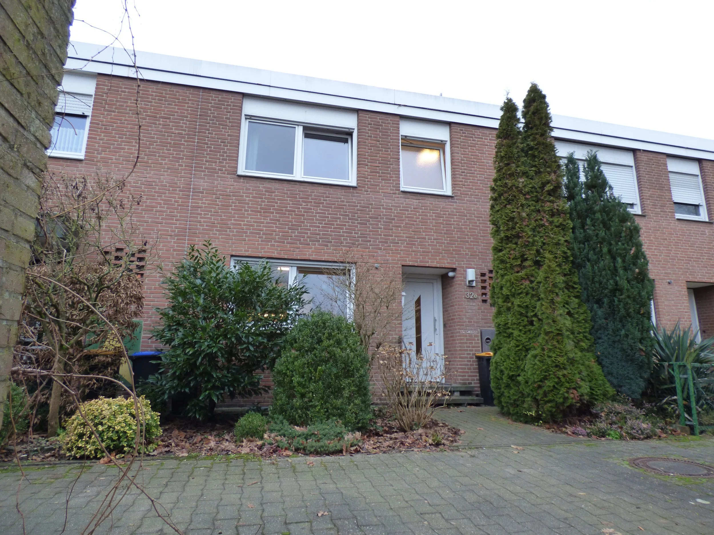 Mehrfamilienhaus mit grauem Eingang, umgeben von Büschen und Bäumen, auf gepflastertem Vorgarten, im Winter oder Herbst ohne Blätter am Baum