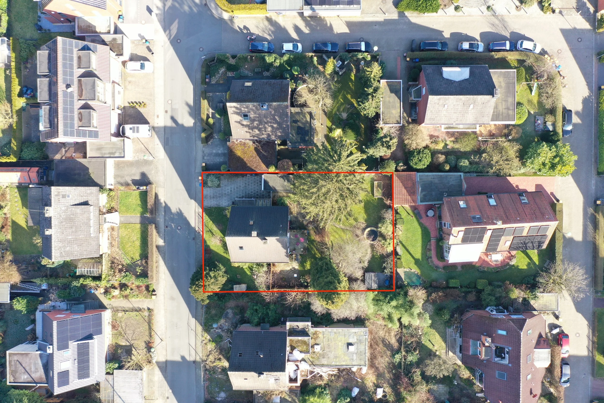 Vogelperspektive auf ein Wohngebiet mit mehreren Häusern, Straßen und Parkplätzen. Im Zentrum ist ein Grundstück mit einem Haus, Bäumen und Garten, umrahmt von einer roten Linie.