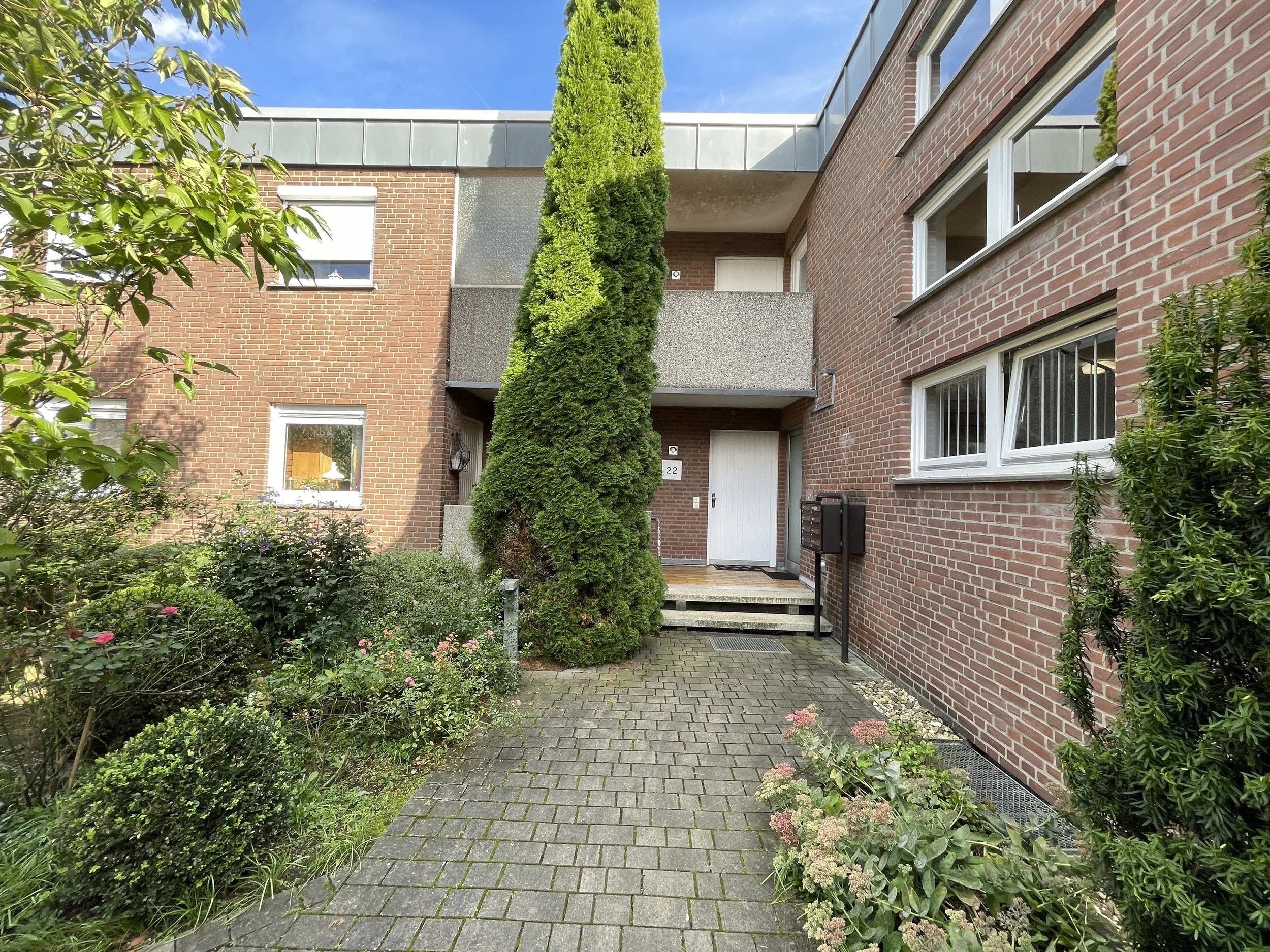 Eingangsbereich eines modernen Mehrfamilienhauses mit roten Ziegelwänden, grünem Garten und einem gepflasterten Weg.