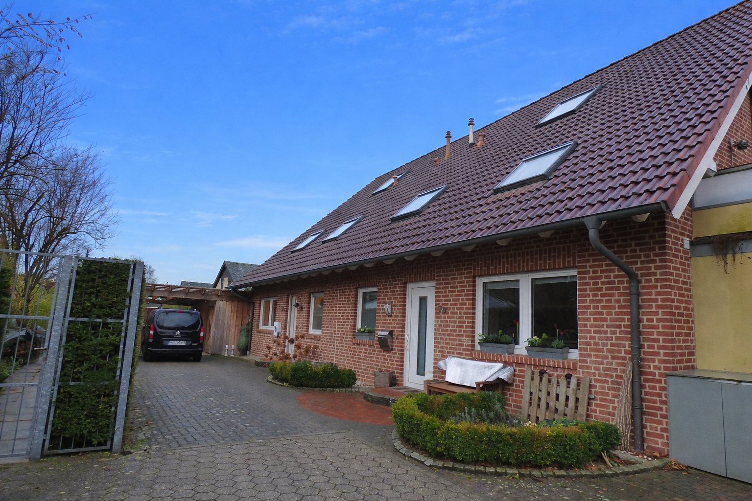 Ein rotes Backsteinhaus mit einem geneigten Dach und mehreren Dachfenstern, umgeben von einem gepflasterten Hof mit kleinen Gartenflächen und Bäumen.