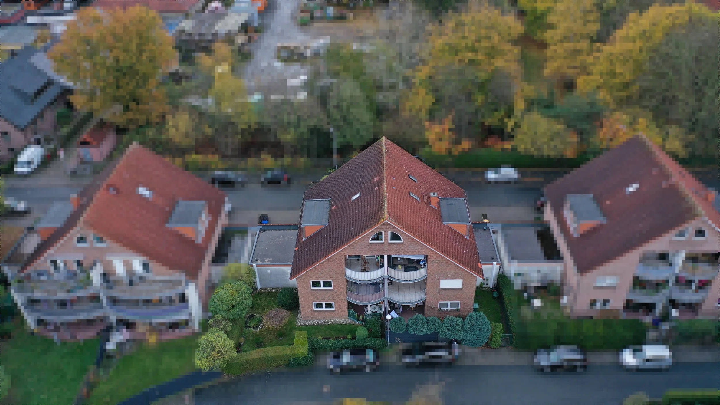 Vogelperspektive auf ein dreistöckiges Wohnhaus mit roten Ziegeldach und Balkon im Vordergrund, umgeben von weiteren Mehrfamilienhäusern und Bäumen im Herbst.