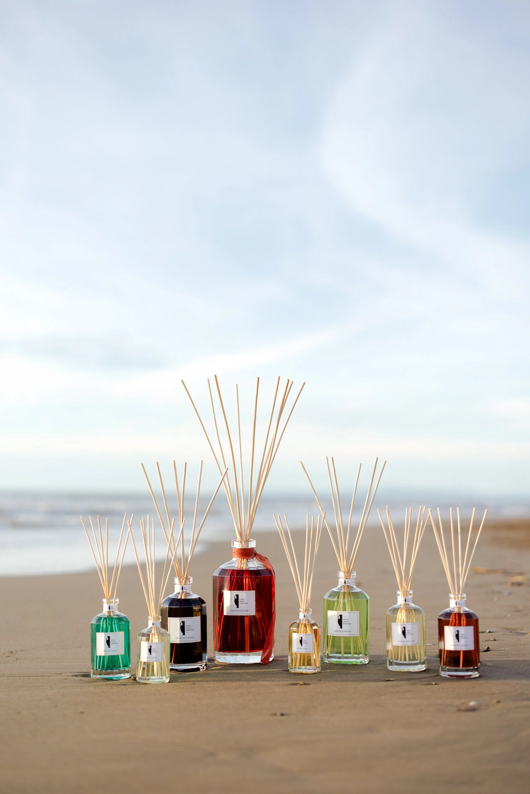 Seven Costa degli Etruschi home fragrances in diffusers with colorful liquids on a sandy beach with the ocean and blue sky in the background.