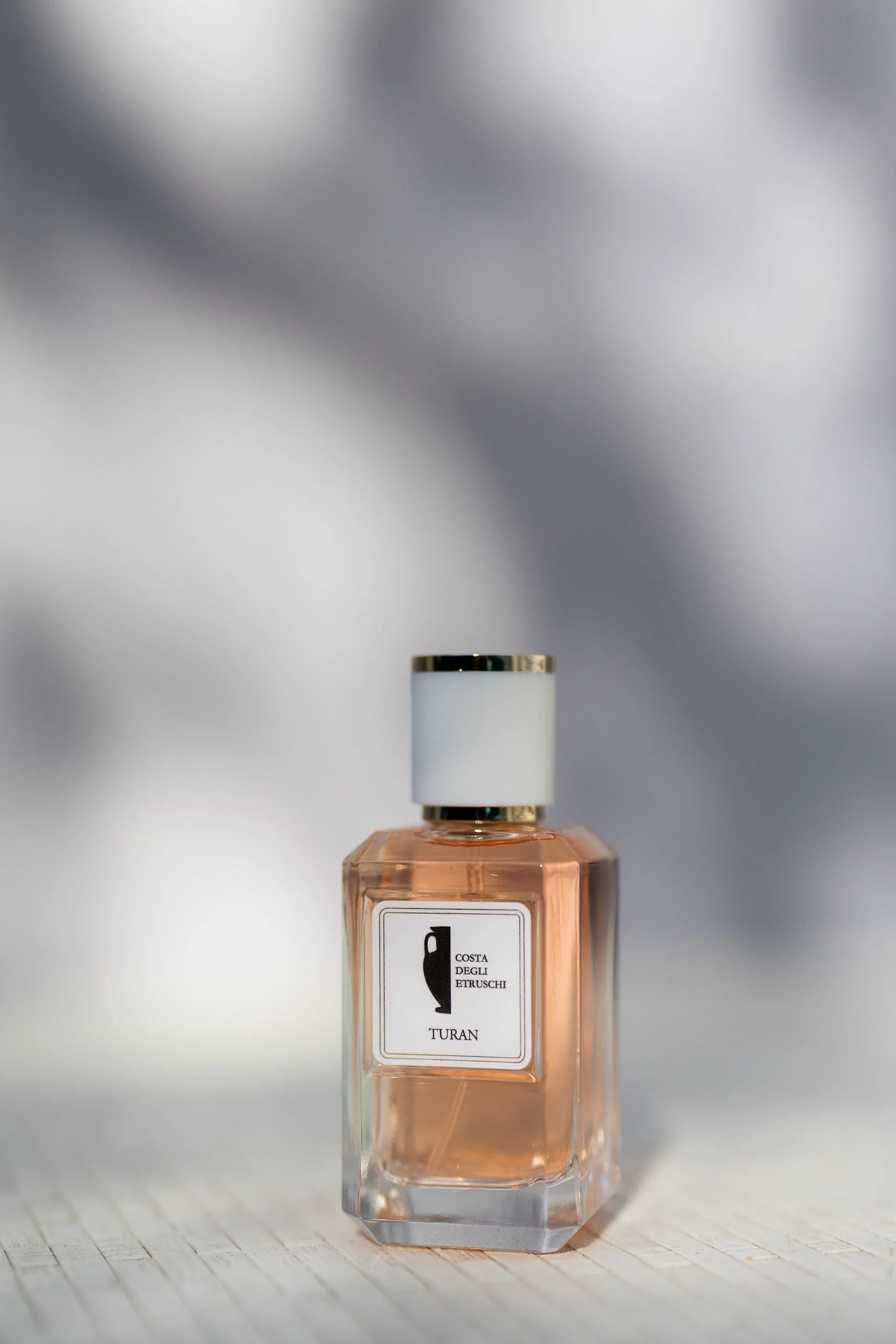 A bottle of perfume with a transparent rectangular glass container, light pink liquid inside, white label, and a white cap with gold trim, placed on a wooden surface against a blurred background.