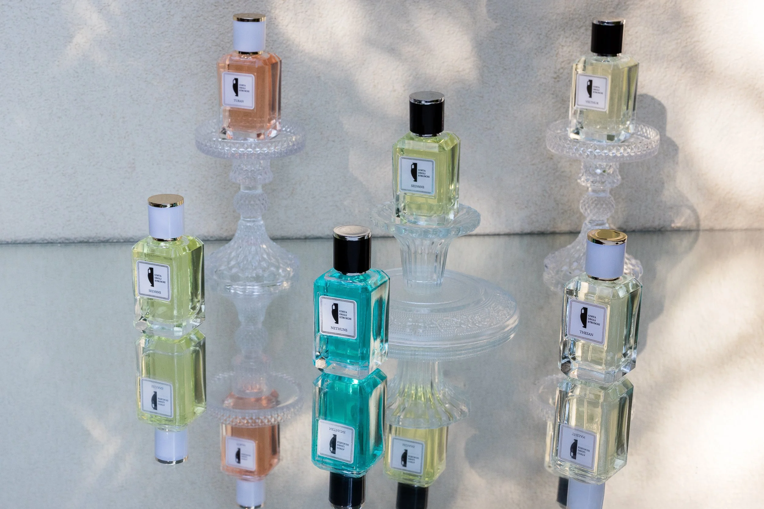 Seven bottles of  Costa degli Etruschi Personal perfume displayed on glass stands and a reflective surface, with shadows cast on a textured wall behind them.