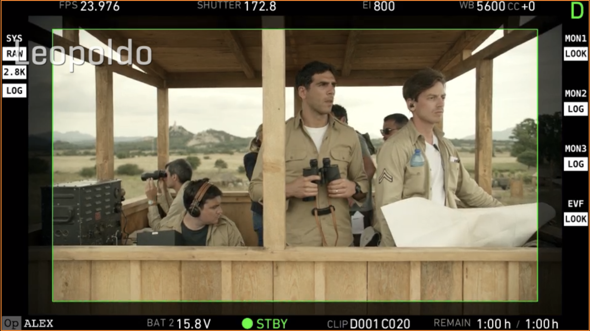Scene from a military operation with four men in beige uniforms inside a wooden observation post overlooking a desert landscape; two men are standing, one holding binoculars and the other a rolled-up paper; two others are seated, one using binoculars and the other with headphones, near a radio or communication device.