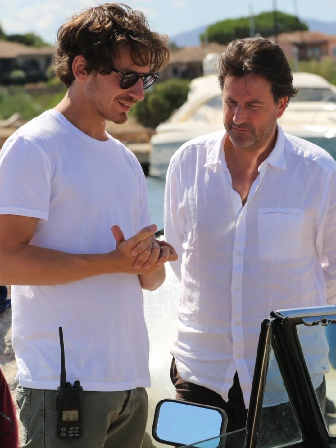 Two men standing outdoors near a marina with boats, one younger with curly hair wearing sunglasses and a white t-shirt, the other older with dark hair wearing a white button-up shirt, looking at a tablet device.