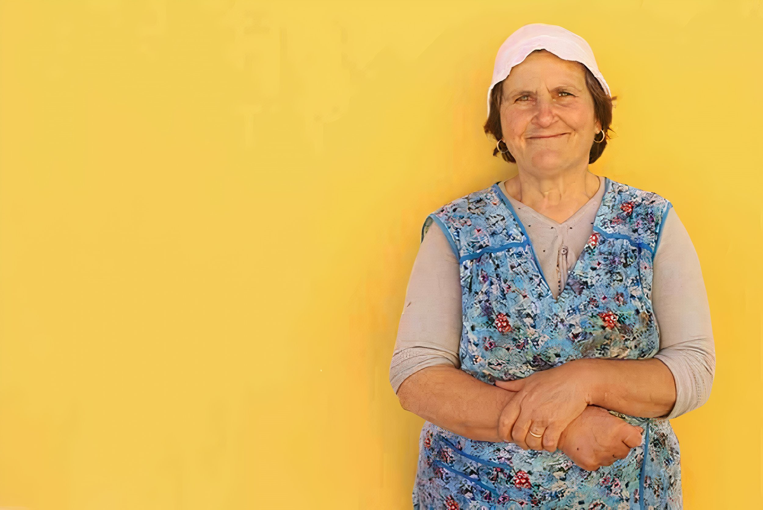 An elderly woman in a pink hat, dressed in a beige T-shirt and a blue apron with floral and geometric patterns, smiles sweetly in front of a yellow background.
