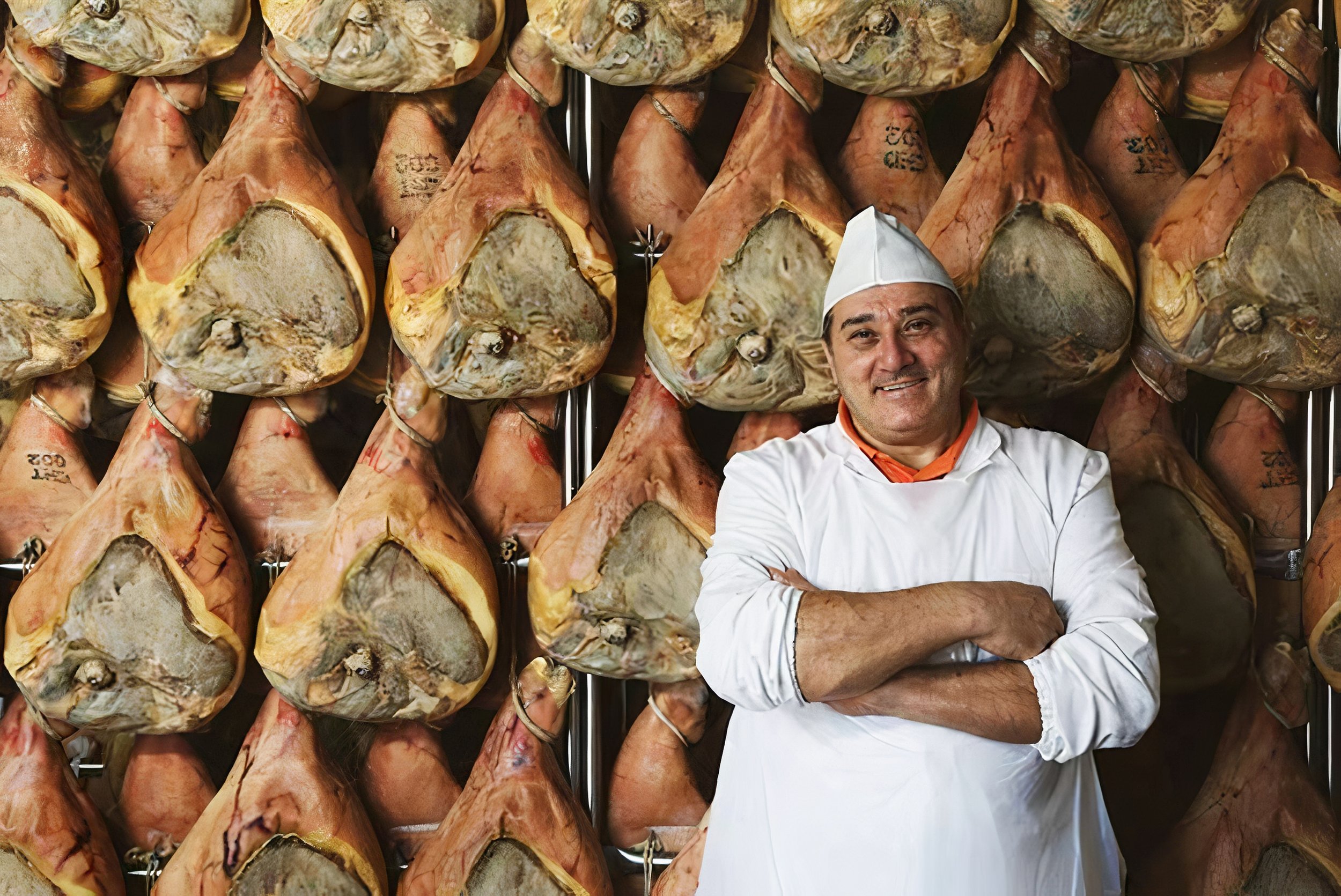 A butcher in a shop with hams hanging in the background.