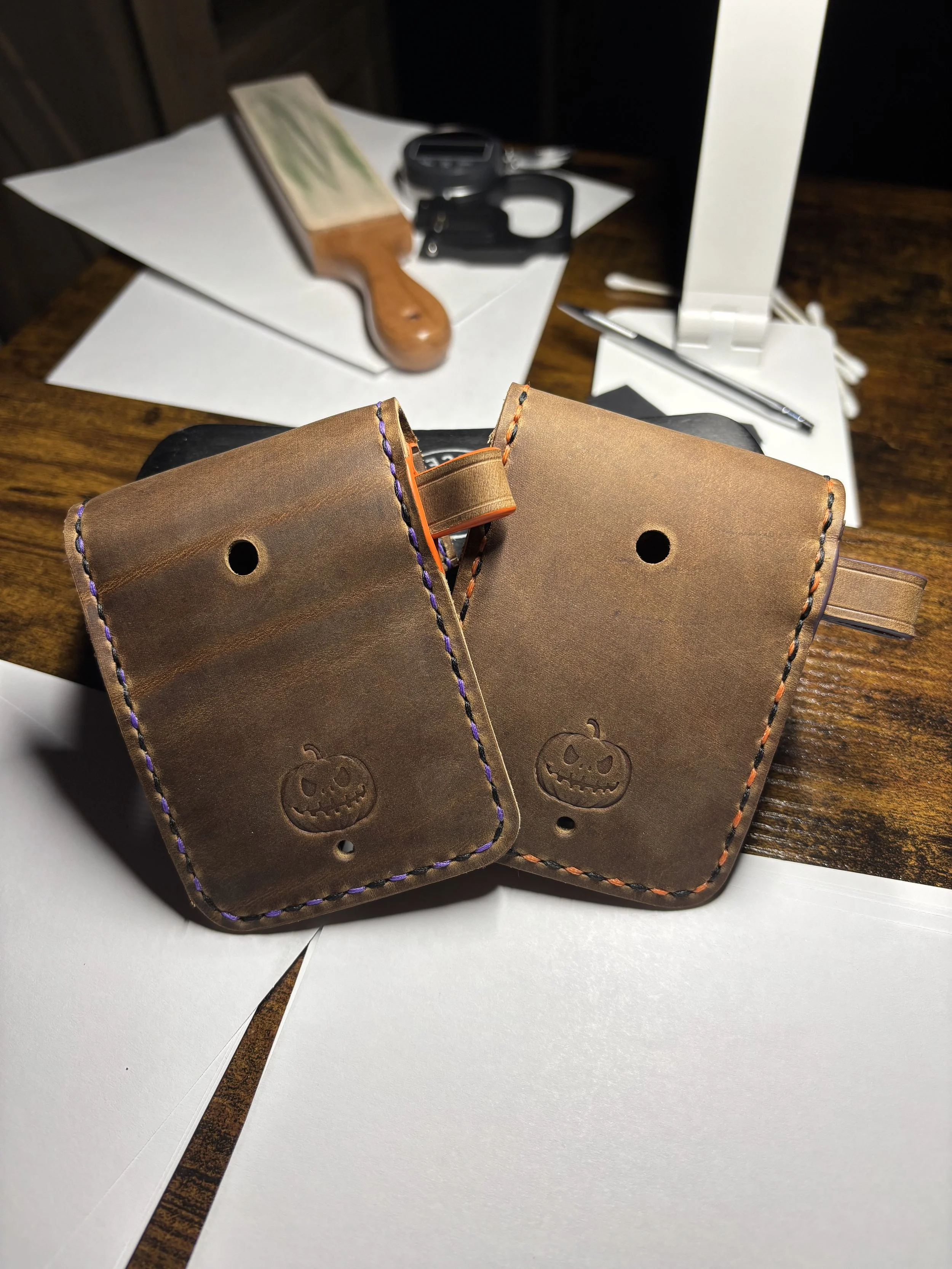 Handcrafted leather Halloween edition dog waste bag holders featuring debossed pumpkin logos and contrasting orange and black; and purple and black saddle stitching on a workbench.