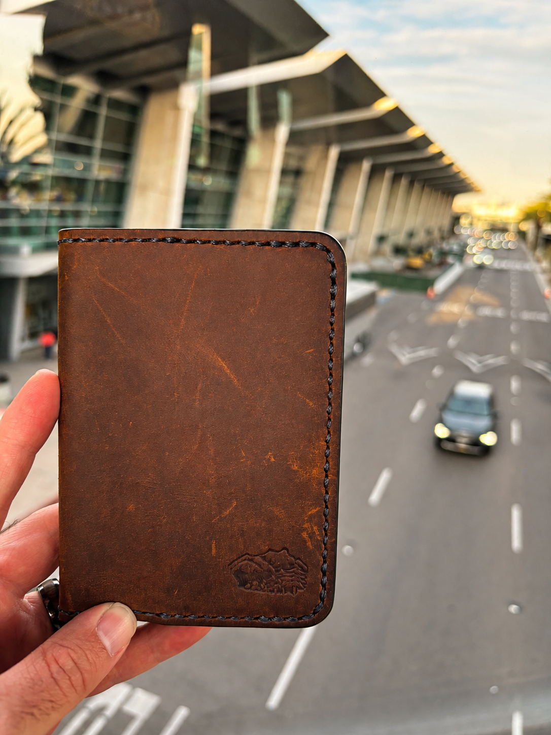 Handheld view of brown leather Rambler passport case with an airport backdrop.