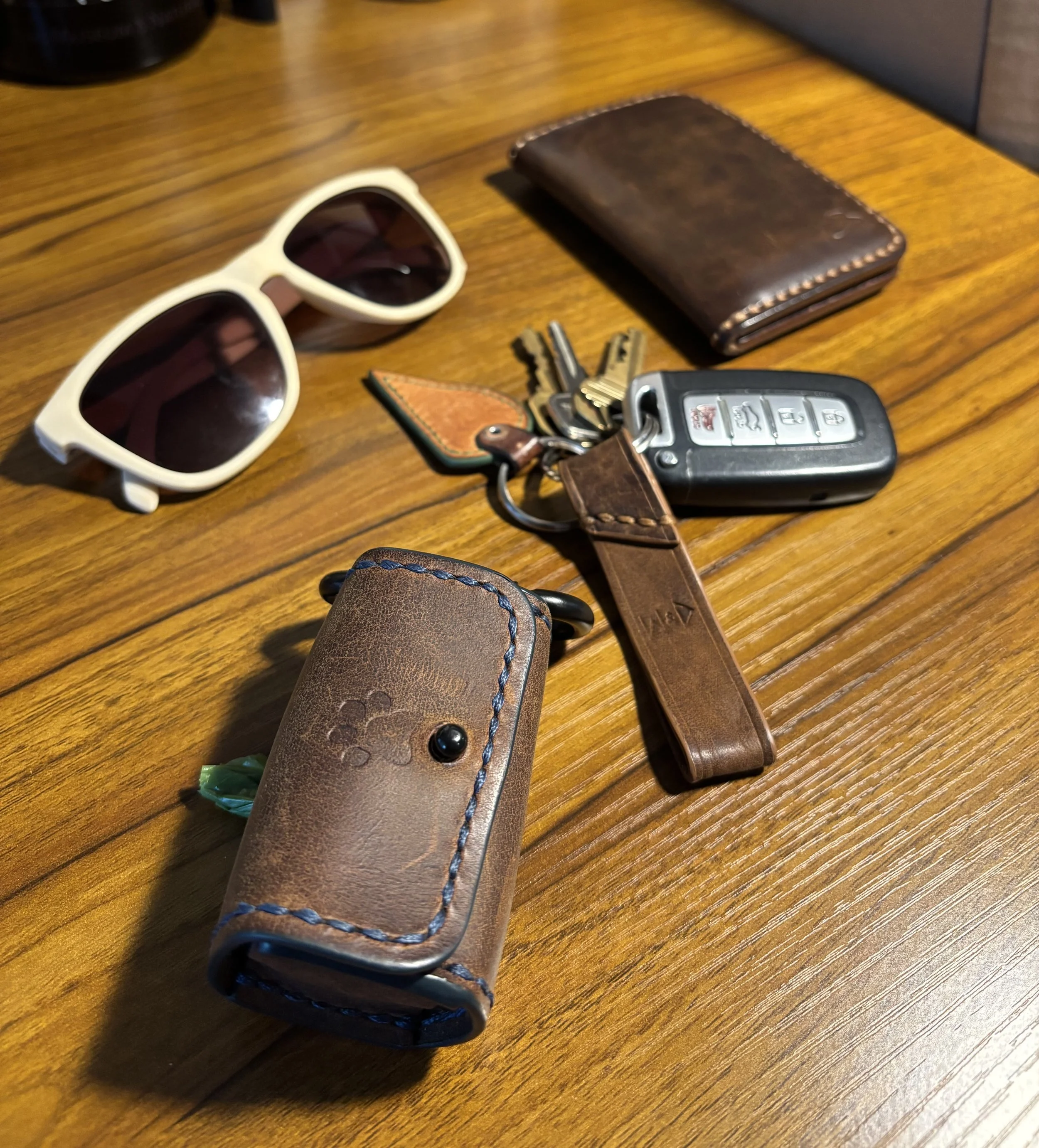 Everyday carry flat lay featuring a brown leather dog waste bag dispenser, leather key fob, bifold wallet, keys, and sunglasses on a wooden table.