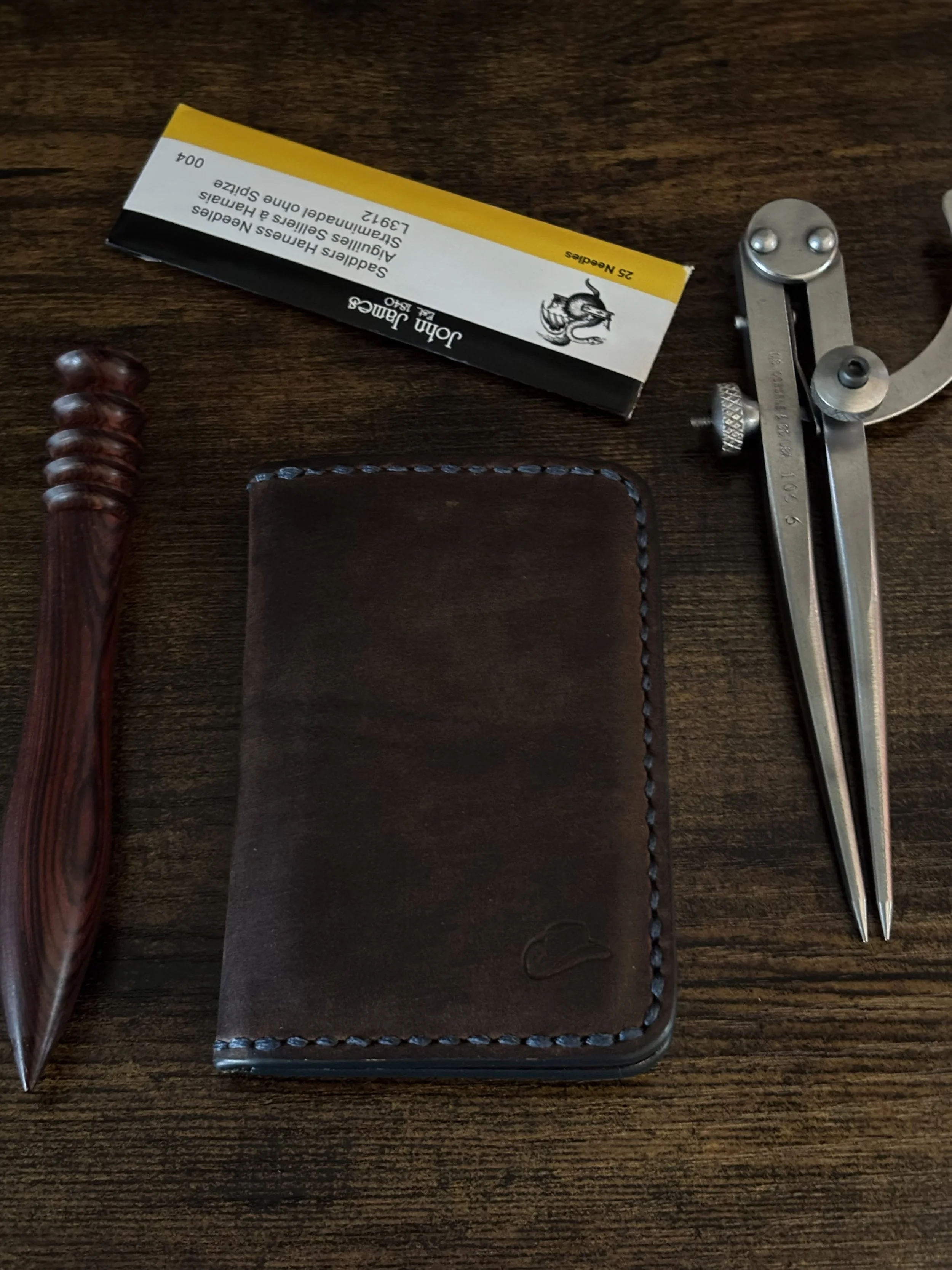 Brown leather bifold wallet arranged with John James needles, wing dividers, and a wood slicker burnisher on a dark workbench.