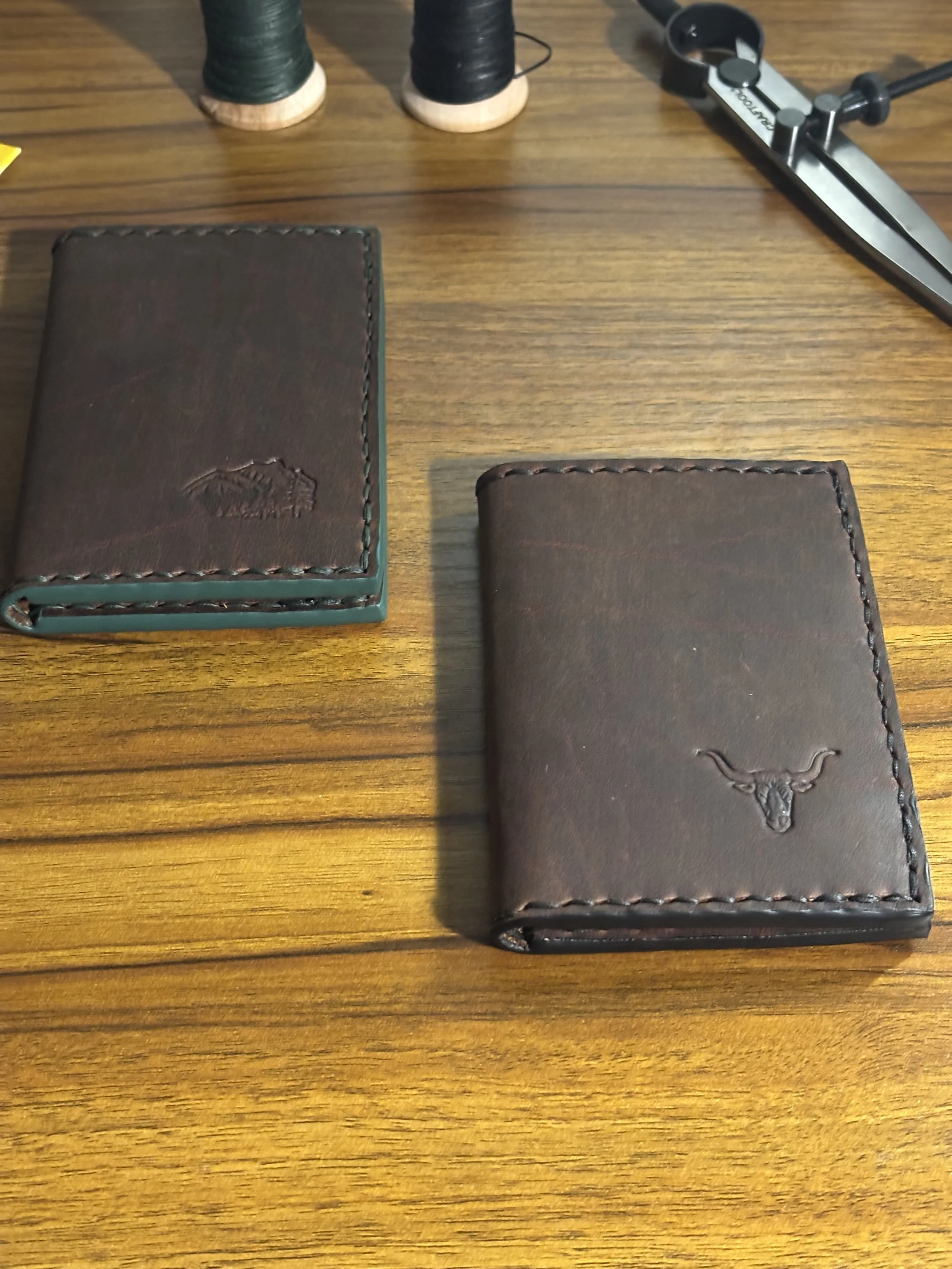Pair of handcrafted brown leather bifold wallets resting side by side on a wooden workbench, highlighting hand painted edges.