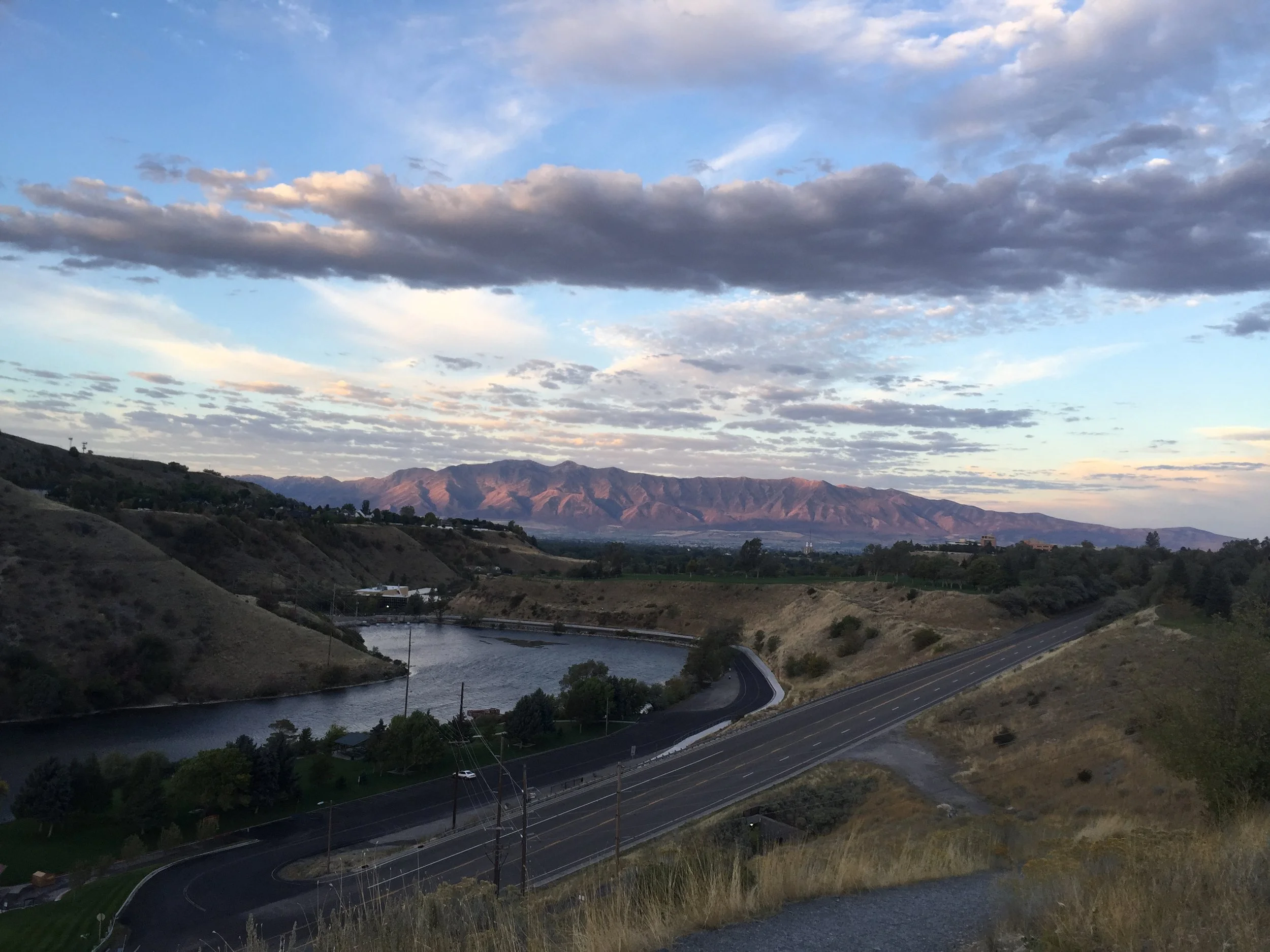 Scenic overlook of Cache Valley, Utah, featuring a curved highway, river reservoir, surrounding hills, and mountains under a clouded sky; taken on trail overlooking first dam and utah state university..