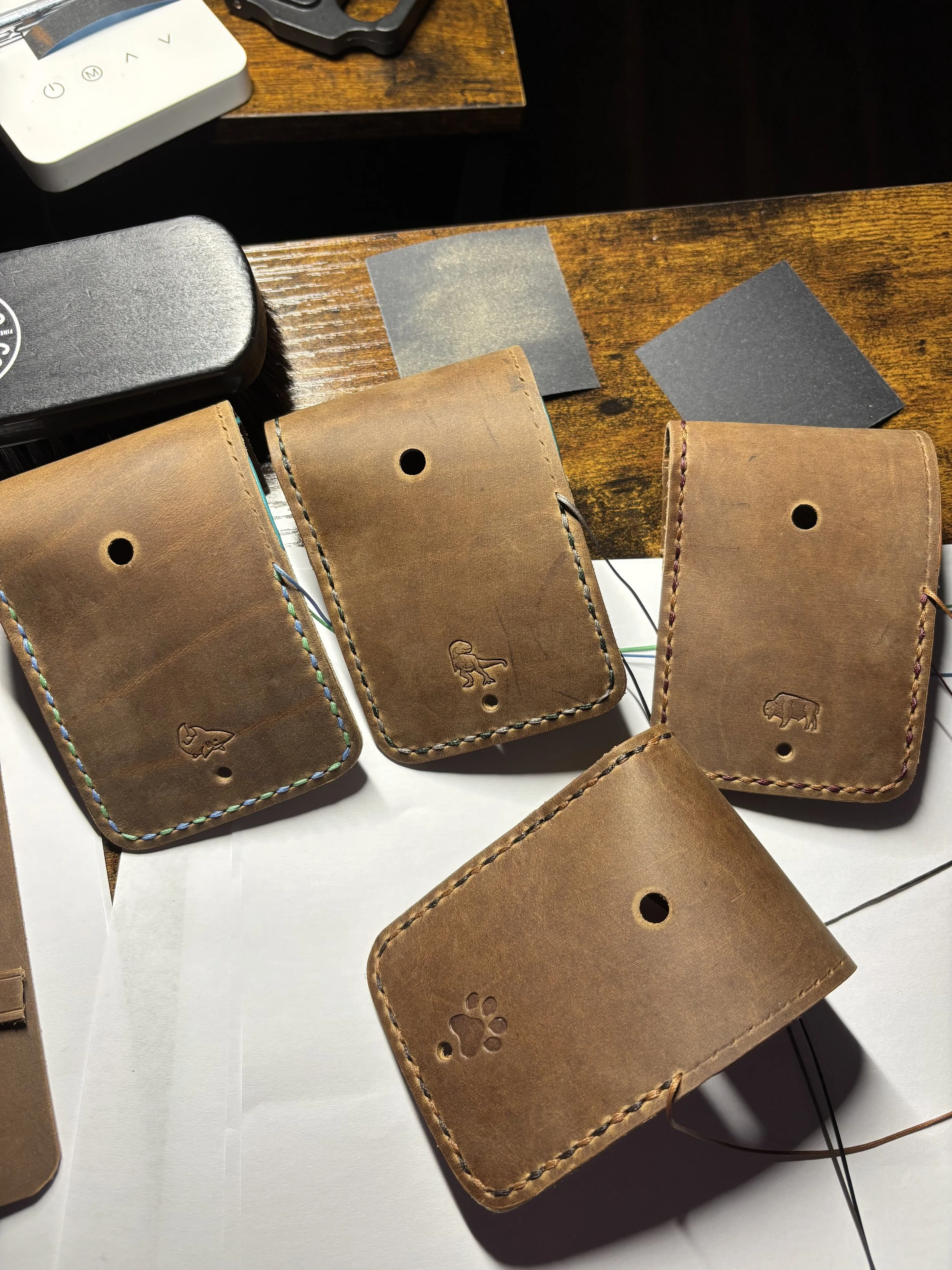 Handcrafted leather dog waste bag holders featuring debossed shark, dinosaur, bison, and paw print designs with dual-colored saddle stitching per piece on a workbench.