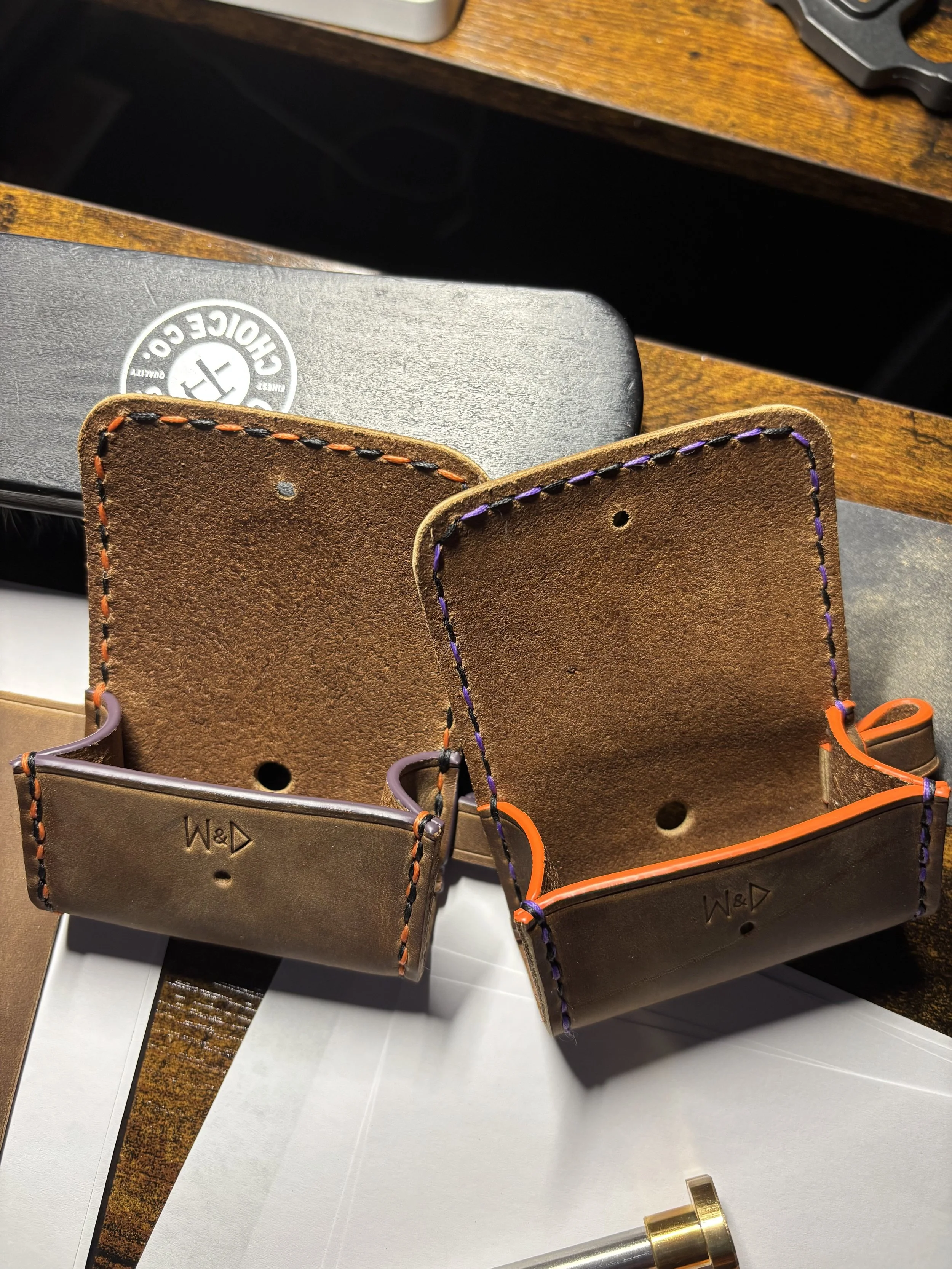 Handcrafted leather Halloween edition dog waste bag holders featuring debossed maker's mark and contrasting orange and black; and purple and black saddle stitching on a workbench