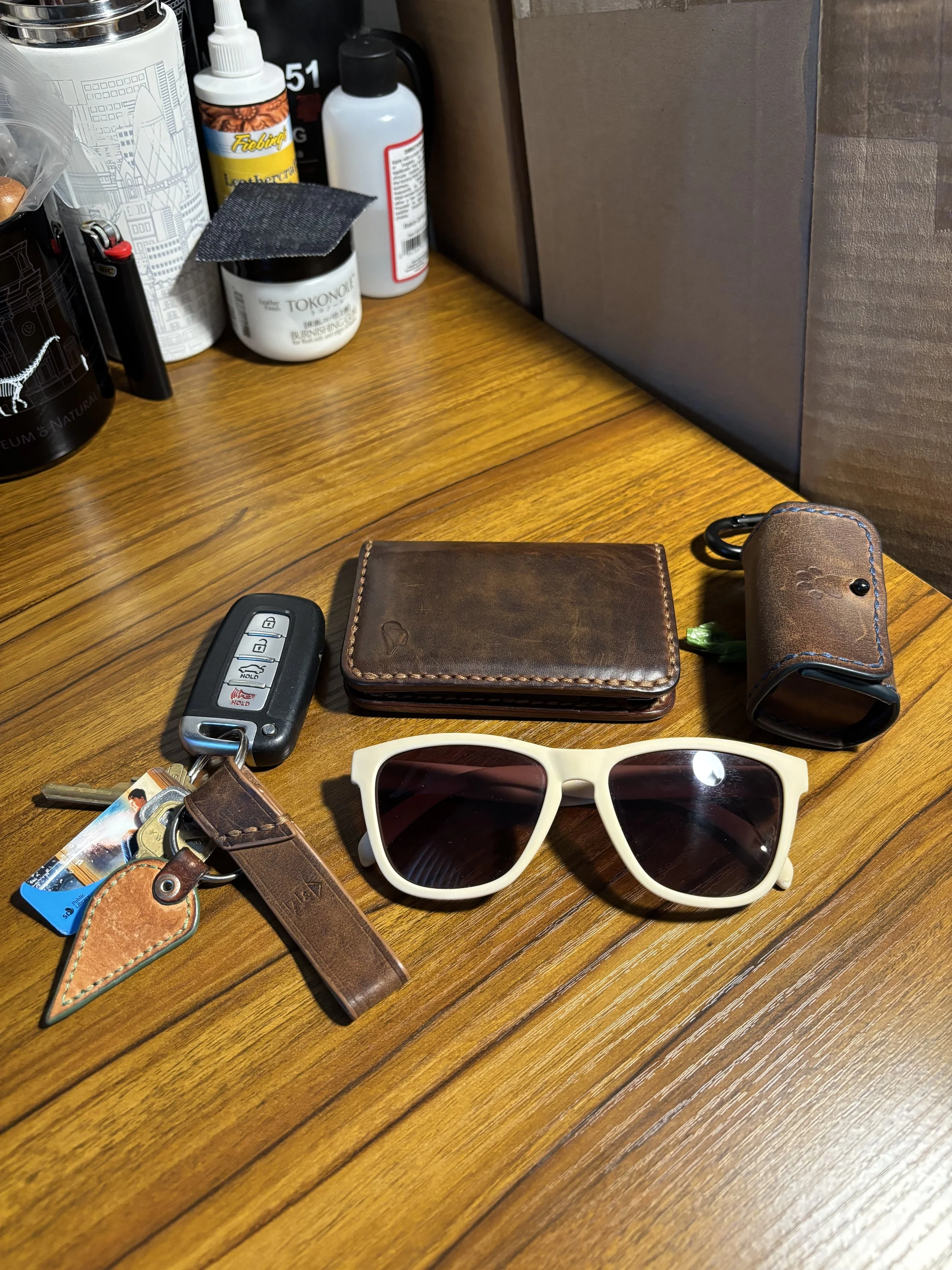 Everyday carry flat lay featuring leather bifold wallet, dog bag holder, key fob, and sunglasses.