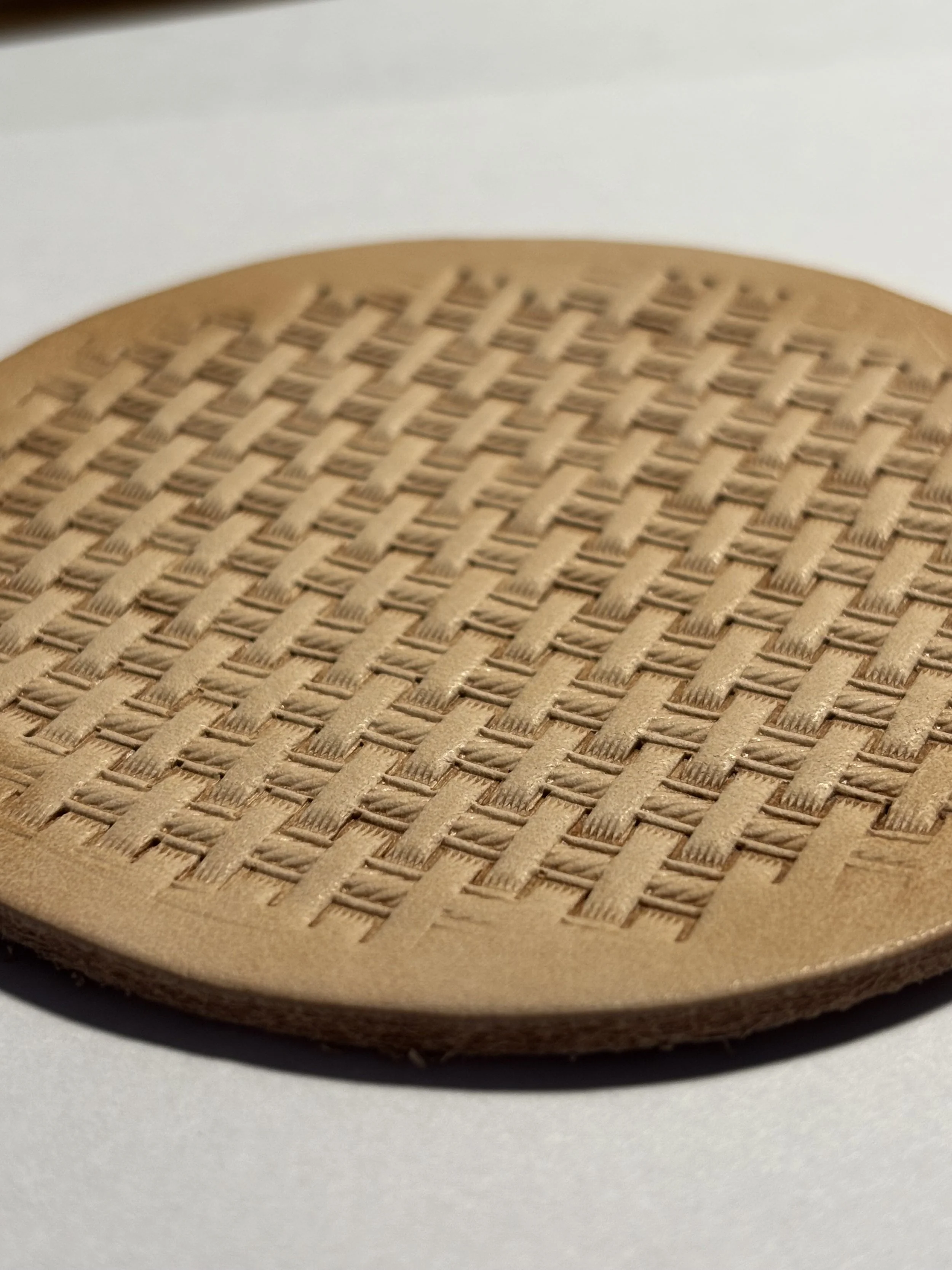 Handmade vegetable-tanned leather coaster featuring a deeply tooled, clean basketweave stamping pattern viewed from a low angle on a light background.