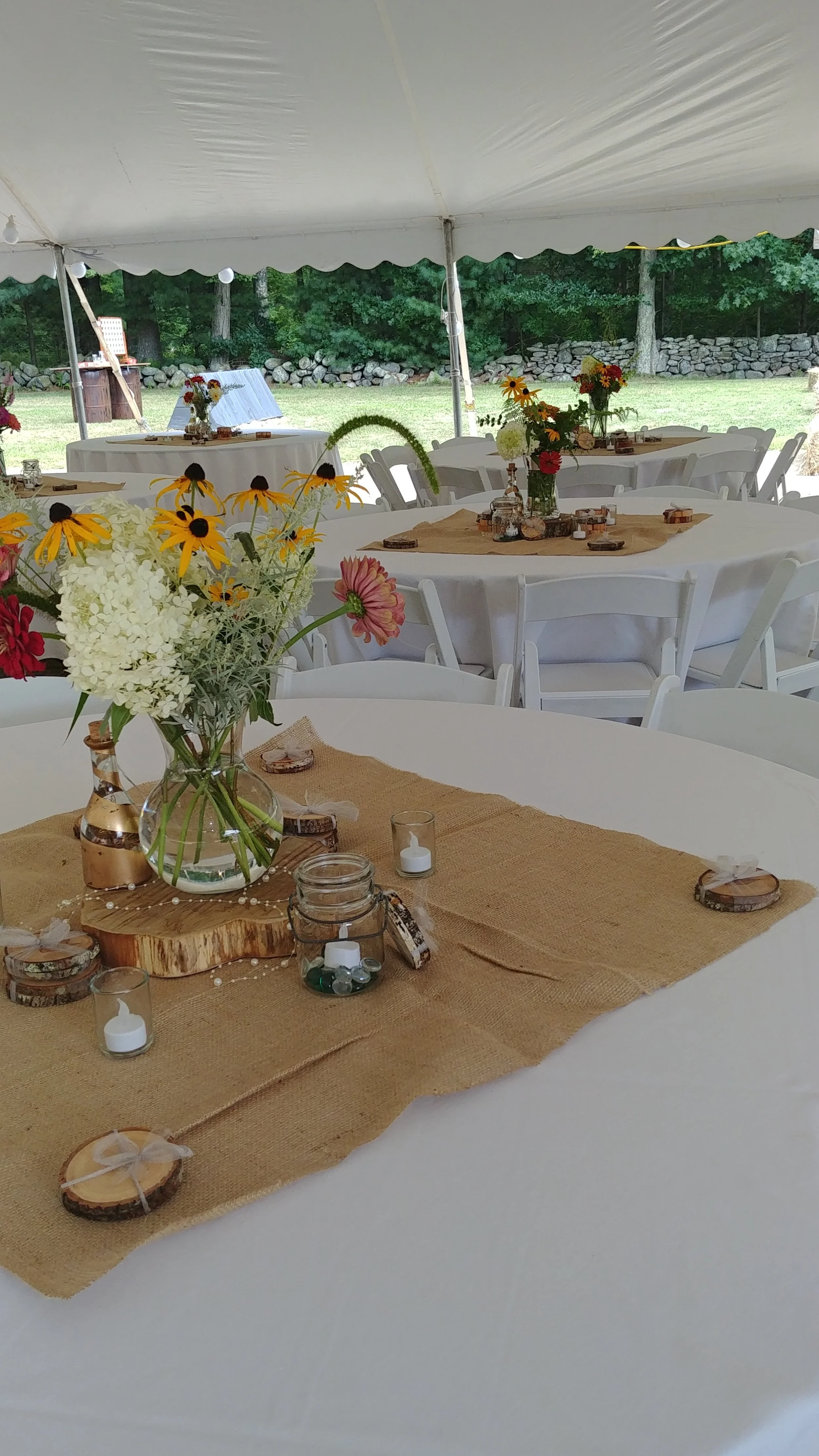 Outdoor wedding reception setup under a tent with round tables decorated with burlap table runners, candles, and flower arrangements in vases, set against a green backyard with trees and stone wall.