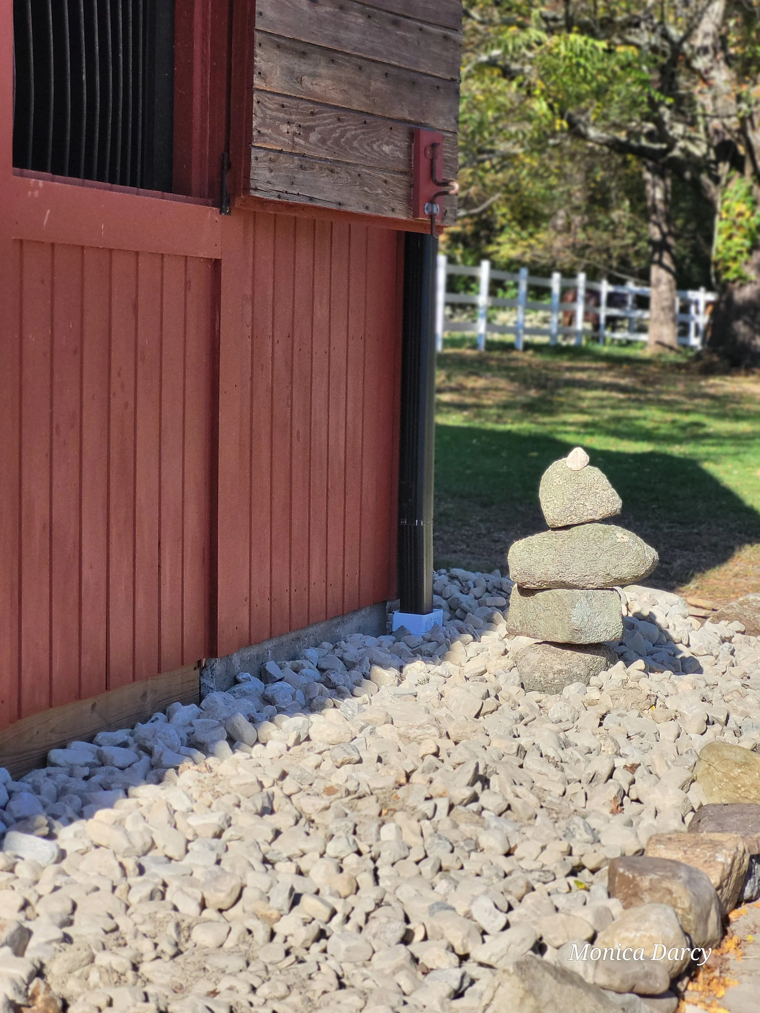A small stack of four carefully balanced stones on a bed of white gravel next to a red wooden building with weathered wood accents and a black downspout, in front of a grassy yard with trees.