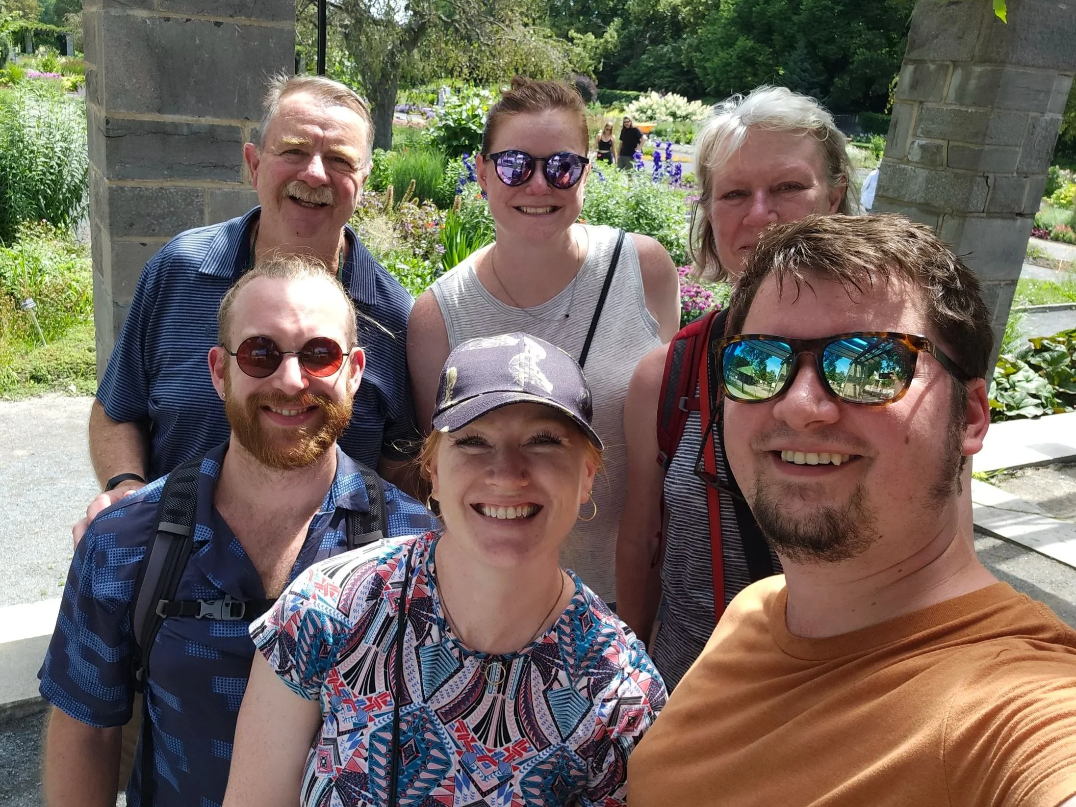 A group of six smiling family members taking a selfie outdoors in a garden with green plants and flowers in the background.