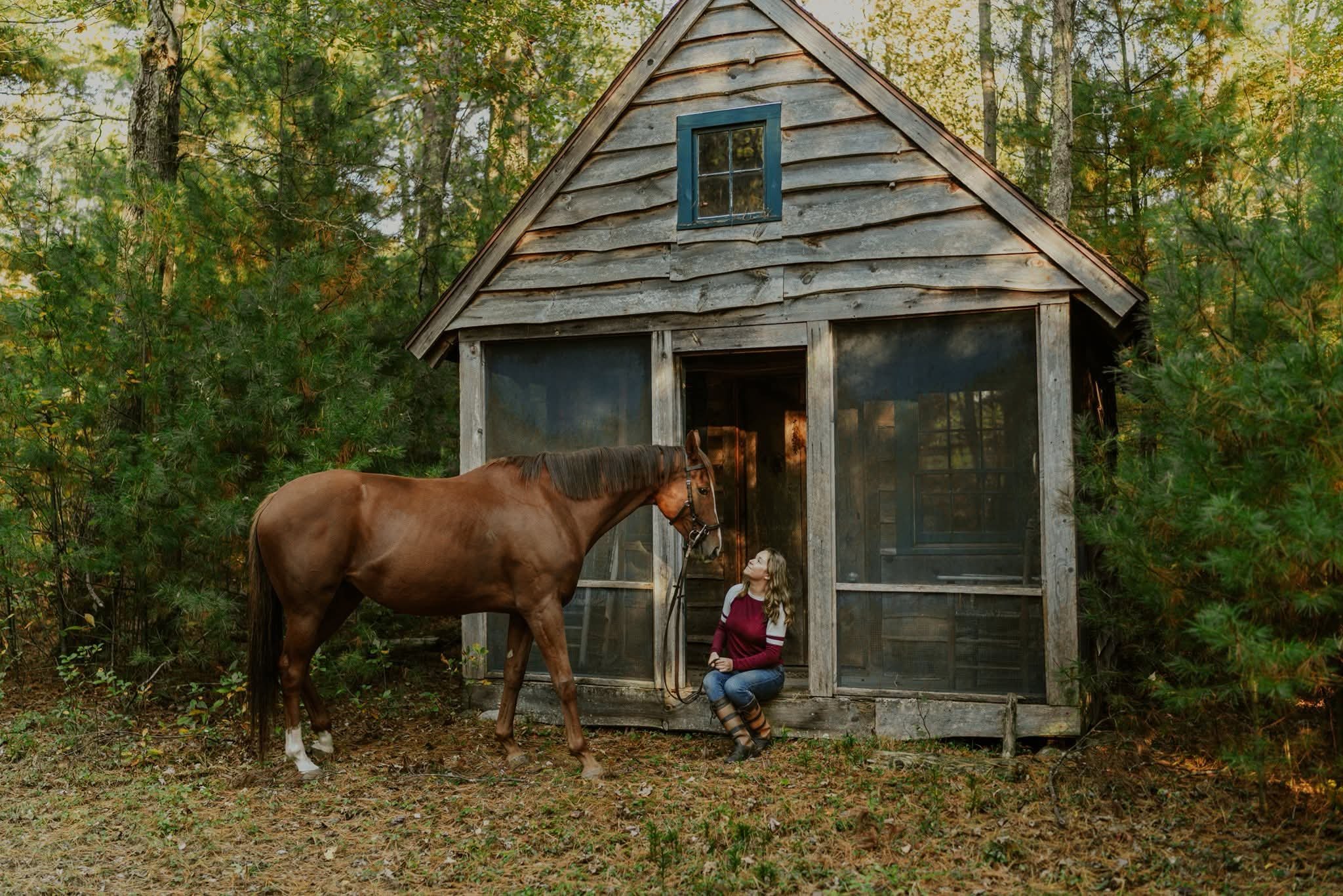 A woman sitting outside a rustic wooden cabin in a wooded area, with a brown horse standing nearby.