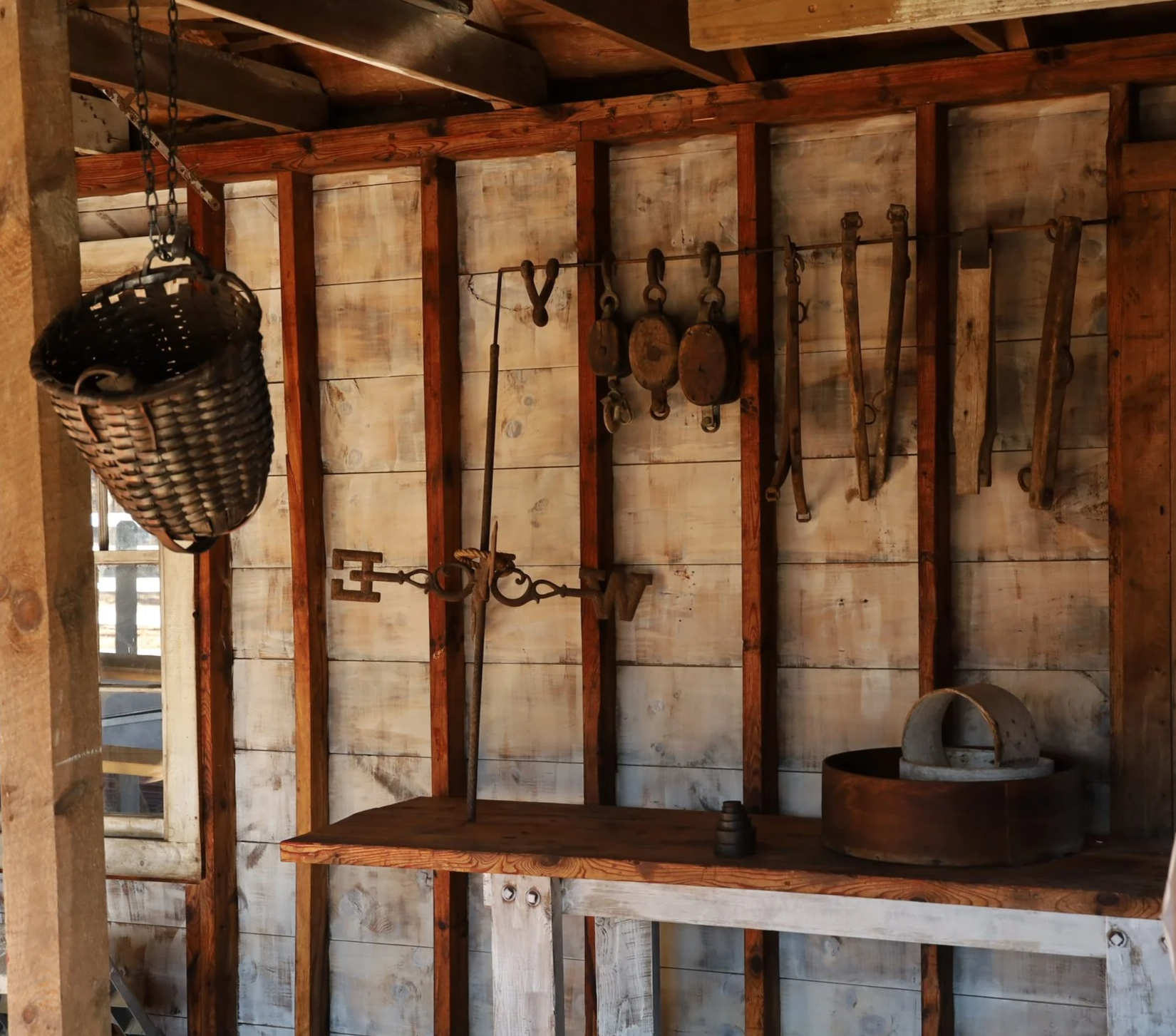 Rustic workshop interior with vintage tools hanging on a wooden wall, including scales, hooks, and a metal bucket.