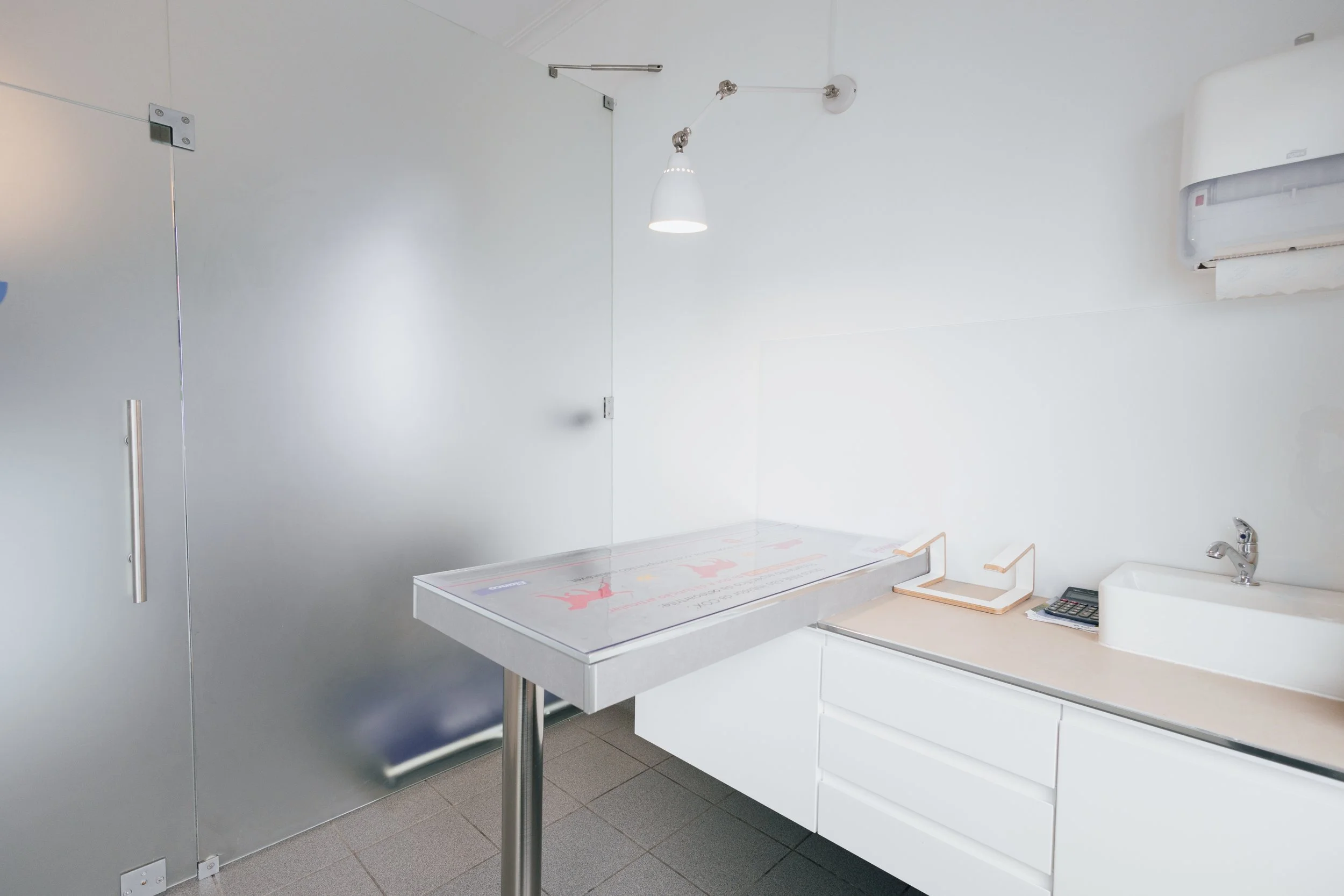 Ambiente com mesa de exame, pia, telefone, controle remoto, lâmpada branca, parede branca e móvel branco.