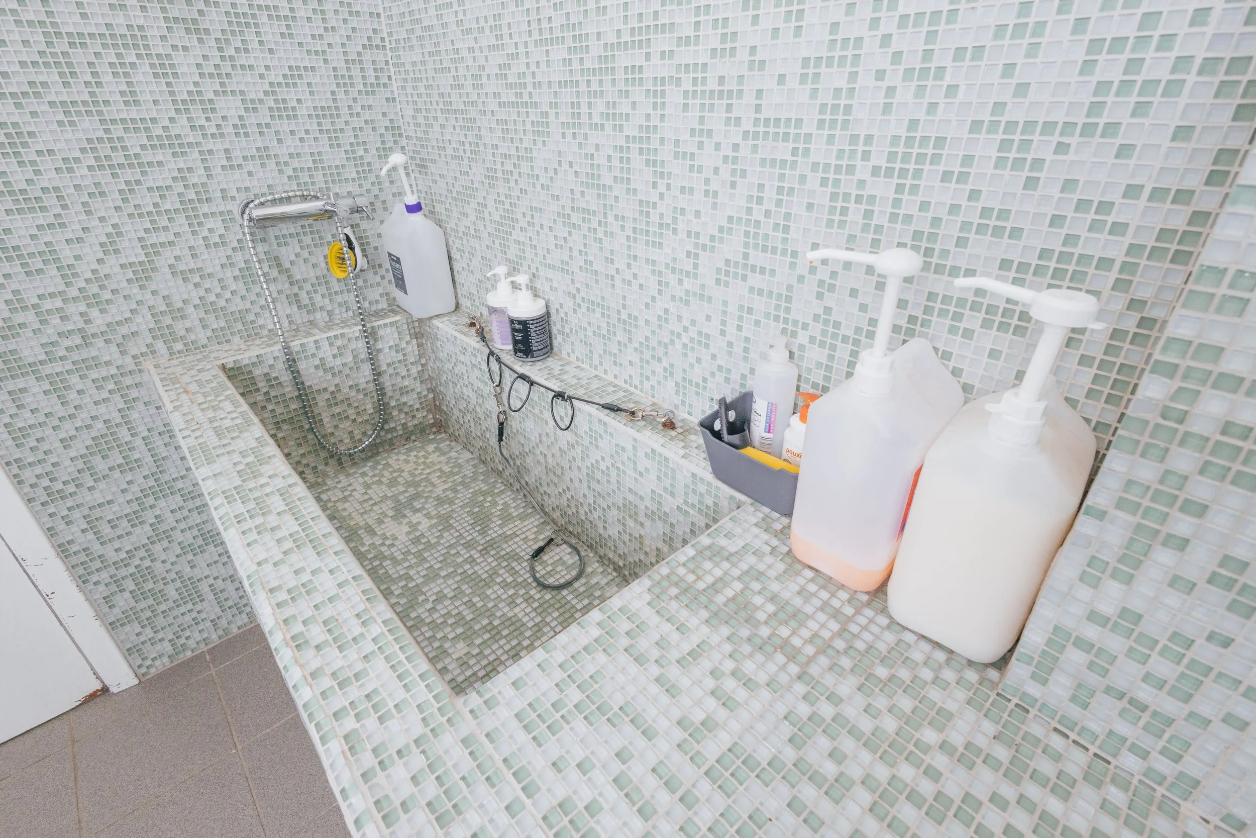 Banheira de mosaico de pedras pequenas em tons de verde, com vários produtos de higiene e ferramentas ao redor, em um ambiente de banho.