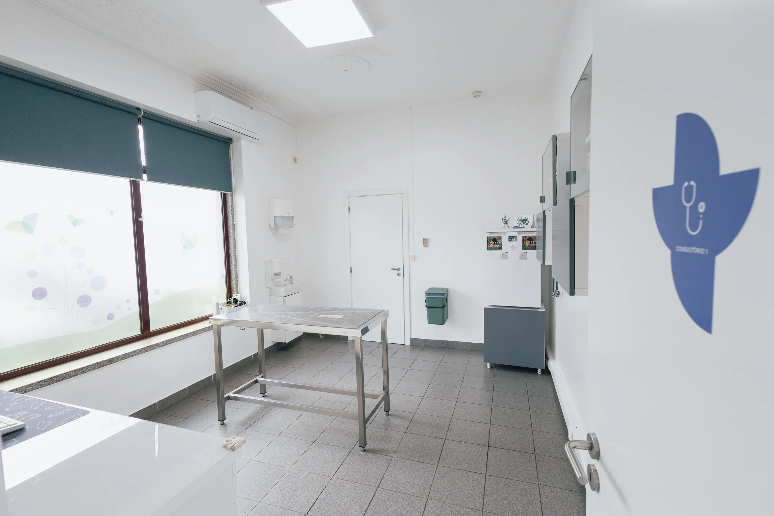 Sala de exame veterinário com mesa de aço, janelas com decoração de animais, geladeira, armários e lixeira.