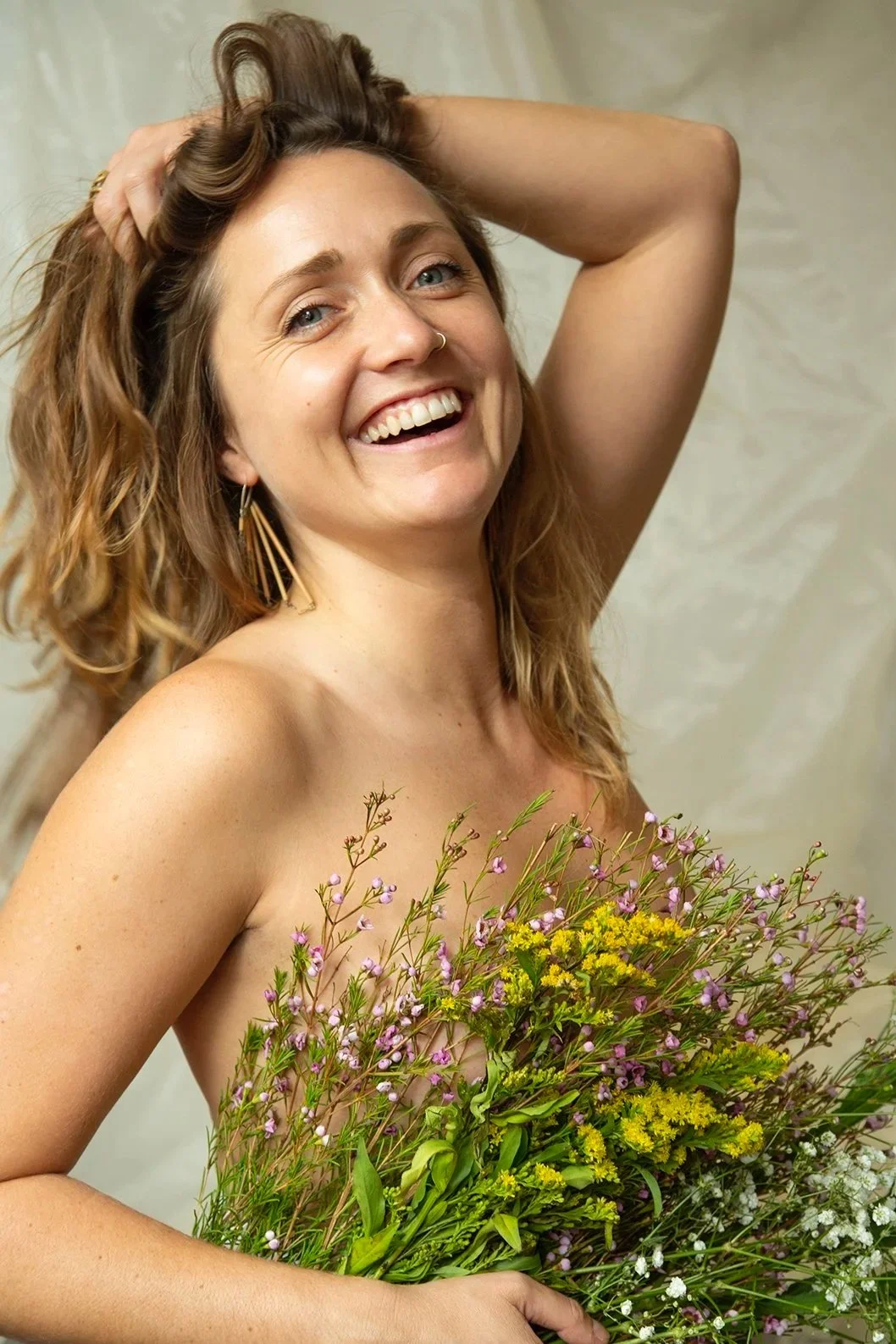 Eine lachende Frau mit lockigem, braunem Haar, hält einen Blumenstrauß mit verschiedenen kleinen, bunten Blumen vor die Brust, während sie eine Hand in den Haaren hat.
