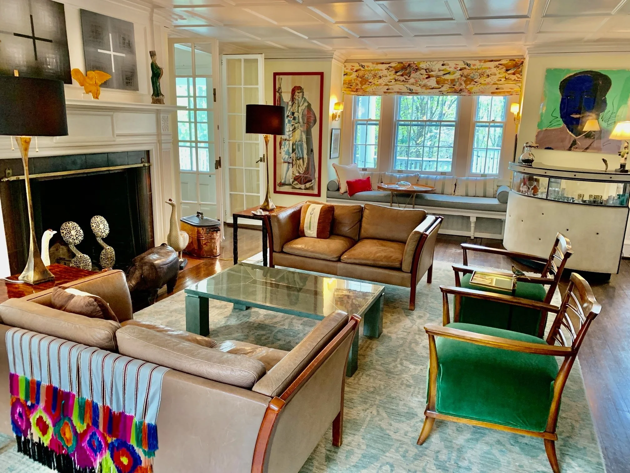 Living room with beige and brown sofas, wooden chairs with green velvet cushions, a glass coffee table, a fireplace with decorative objects, colorful artwork, windows with patterned curtains, and a window seat with striped cushions.