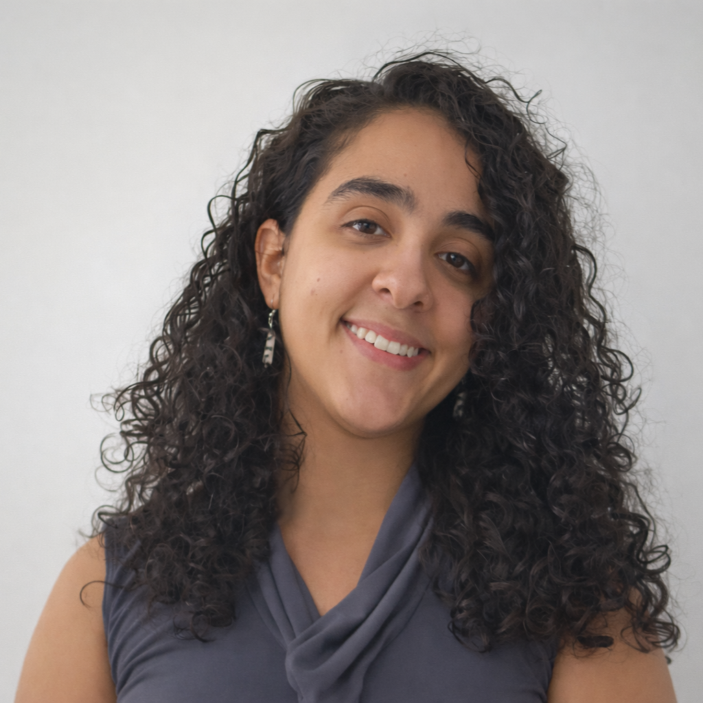 A woman with shoulder-length curly dark hair smiling at the camera, wearing a sleeveless gray top and earrings, standing against a plain white wall.