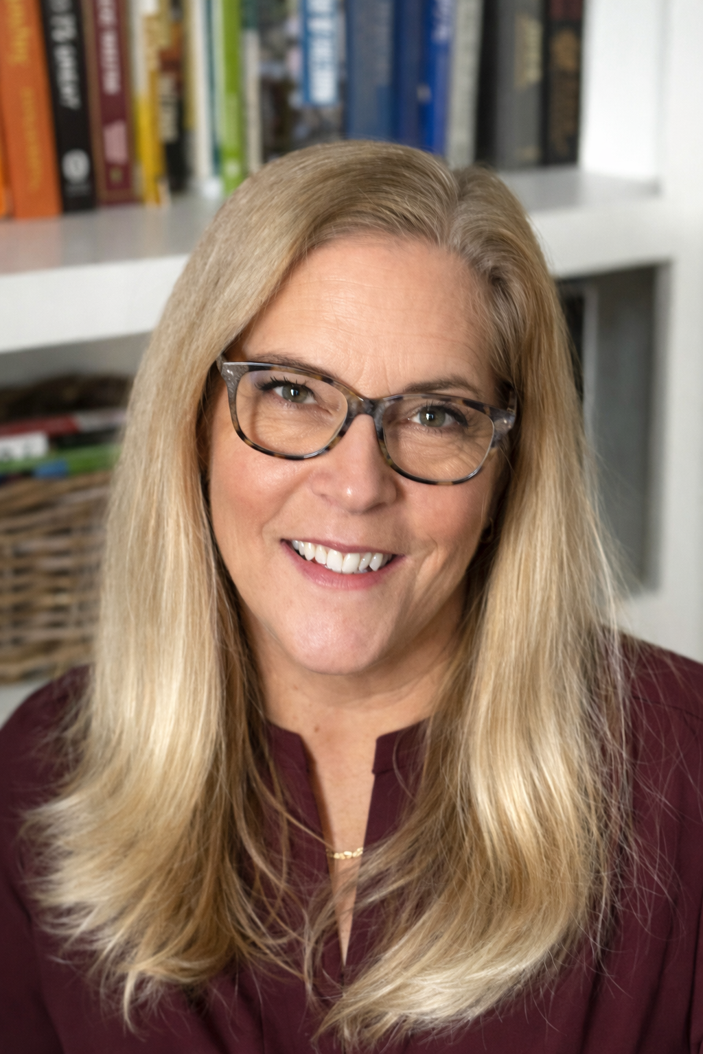 A middle-aged woman with long blonde hair, glasses, and a maroon top smiling in front of a bookshelf filled with books and a woven basket.