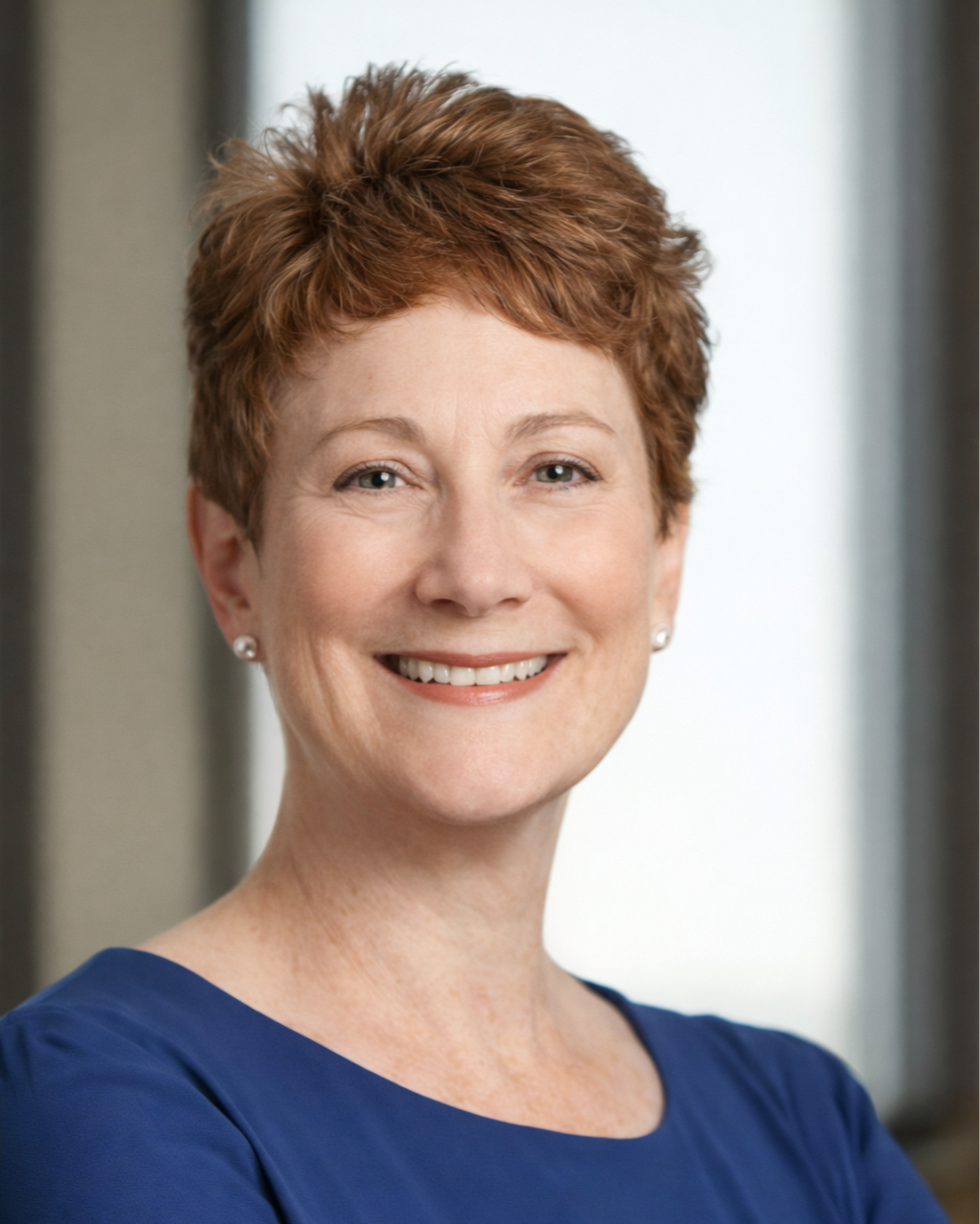 Smiling woman with short hair wearing a blue top, sitting with arms crossed in a modern office setting.