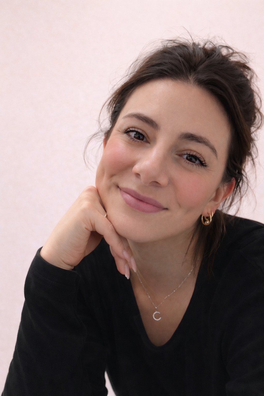 Close-up of a young woman with short brown hair, pink cheeks, and gold jewelry smiling softly, resting her chin on her hand.