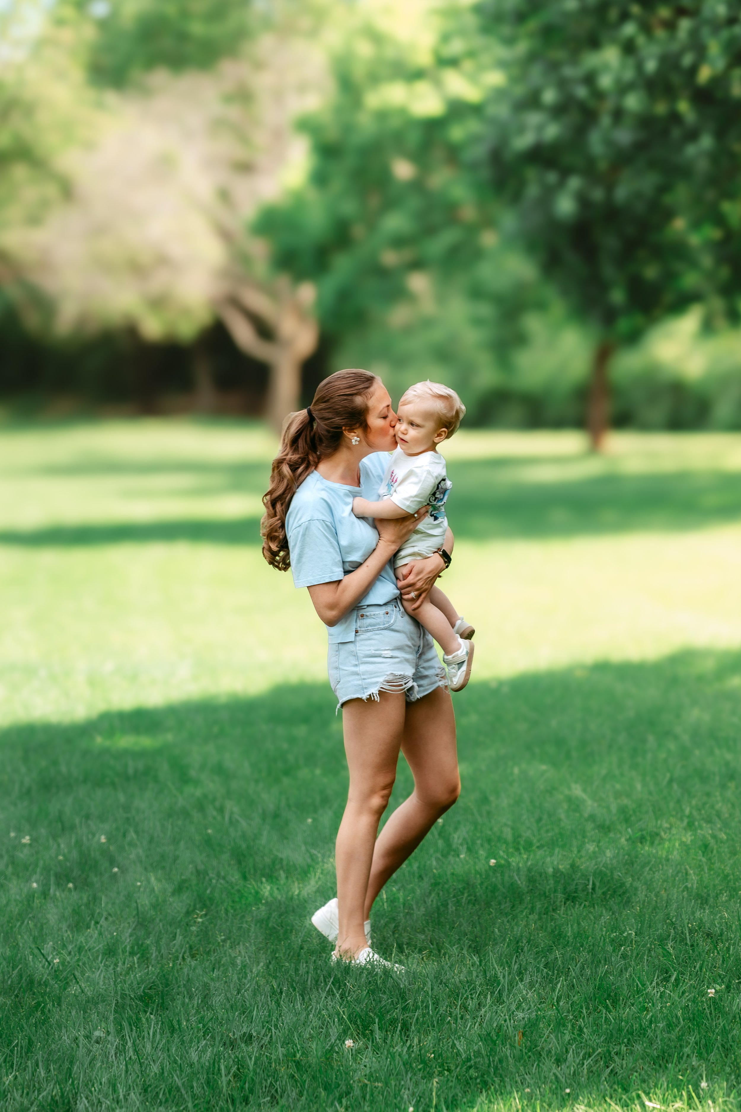 Mother holding and kissing her toddler during a light and airy family photo session in a sunny Gaithersburg, Maryland park.