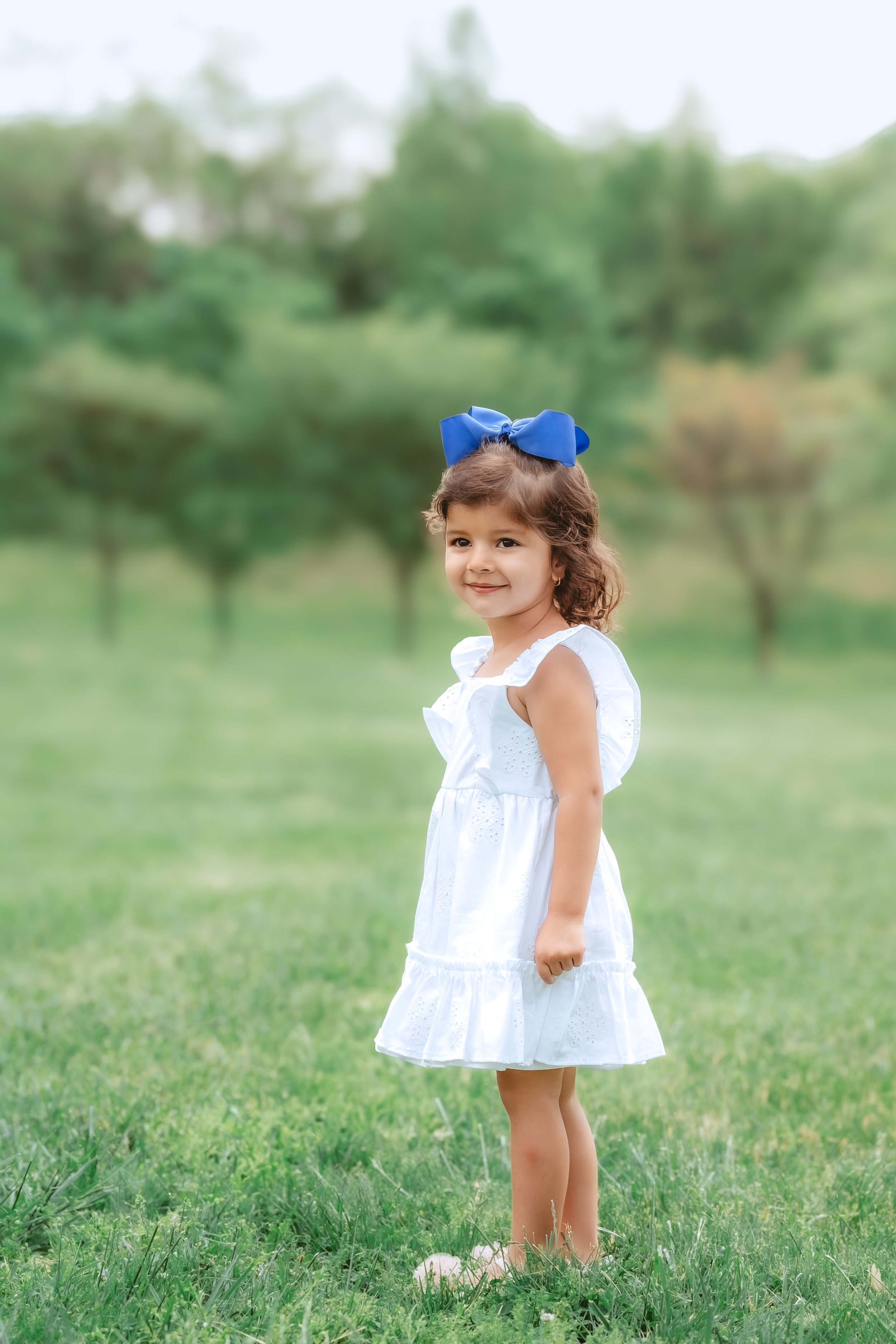 Young girl in a white dress with a blue bow standing in a grassy field during a light and airy family photo session in Gaithersburg, Maryland.