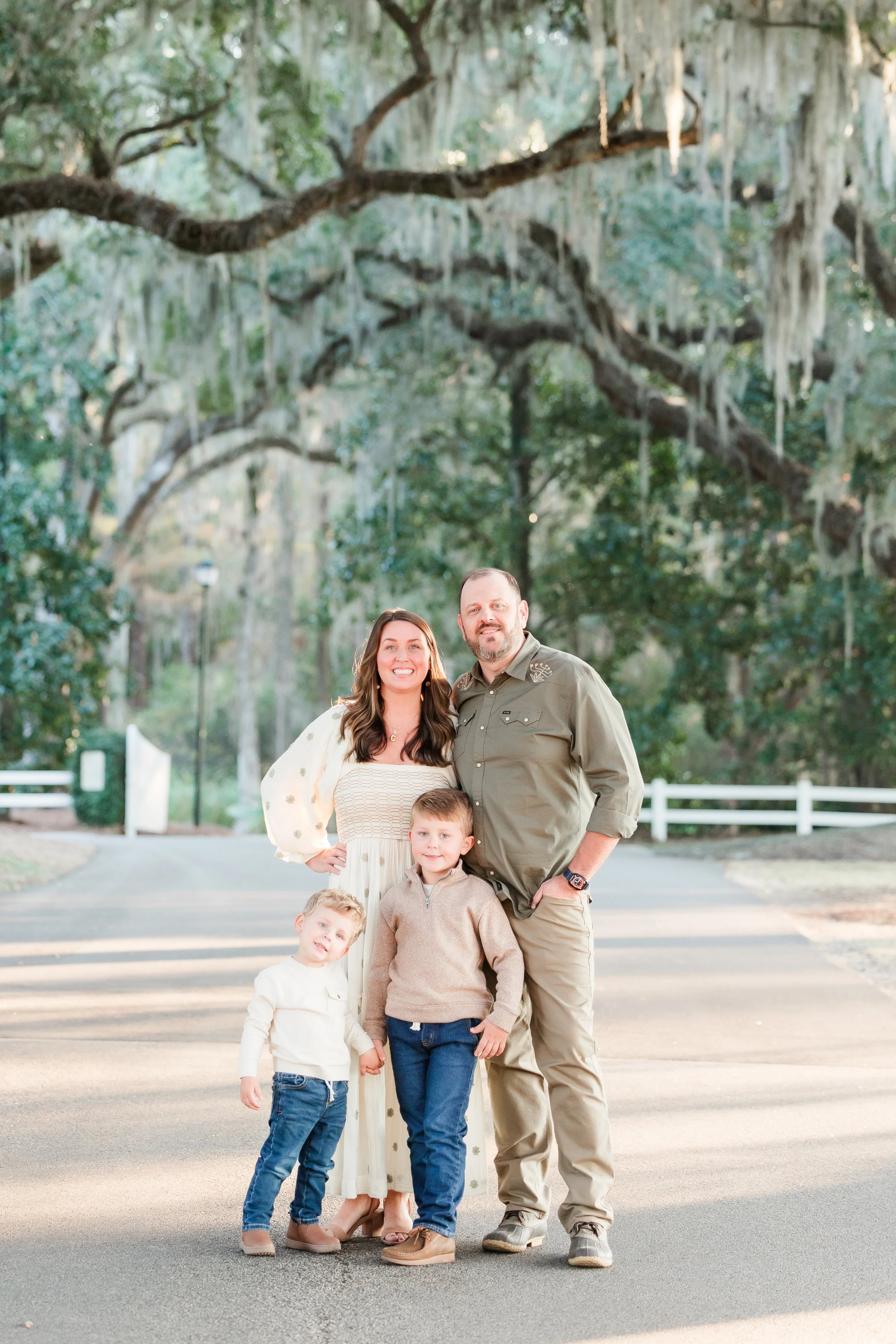 A family of four, two children, a woman, and a man, standing outdoors on a paved path with large trees with hanging moss in the background at Caledonia in Pawleys Island, SC, all smiling at the camera.