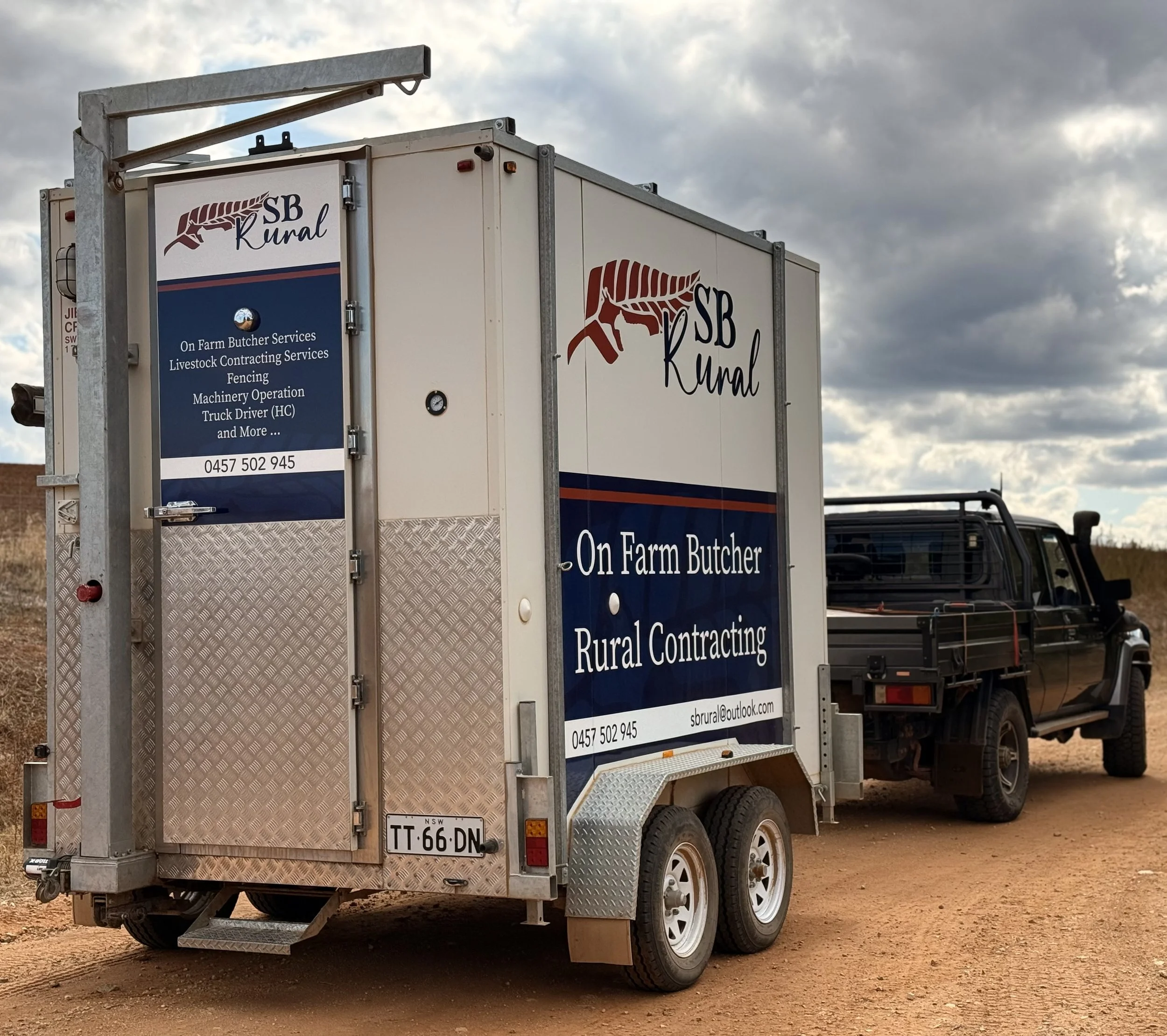 A mobile trailer with branding for SB Rural, a company offering on-farm butcher services and rural contracting, attached to a black pickup truck parked on an unpaved dirt surface under a cloudy sky.