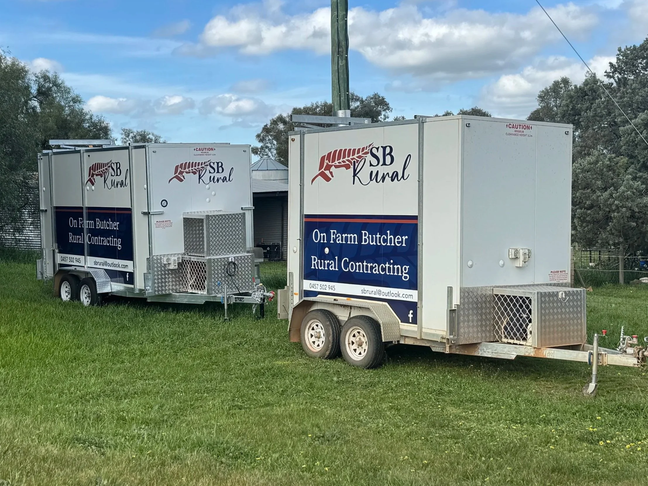 Two white Cool room trailers with signage for SB Rural, a farm butcher and rural contracting business, parked on a grassy field under a partly cloudy sky.