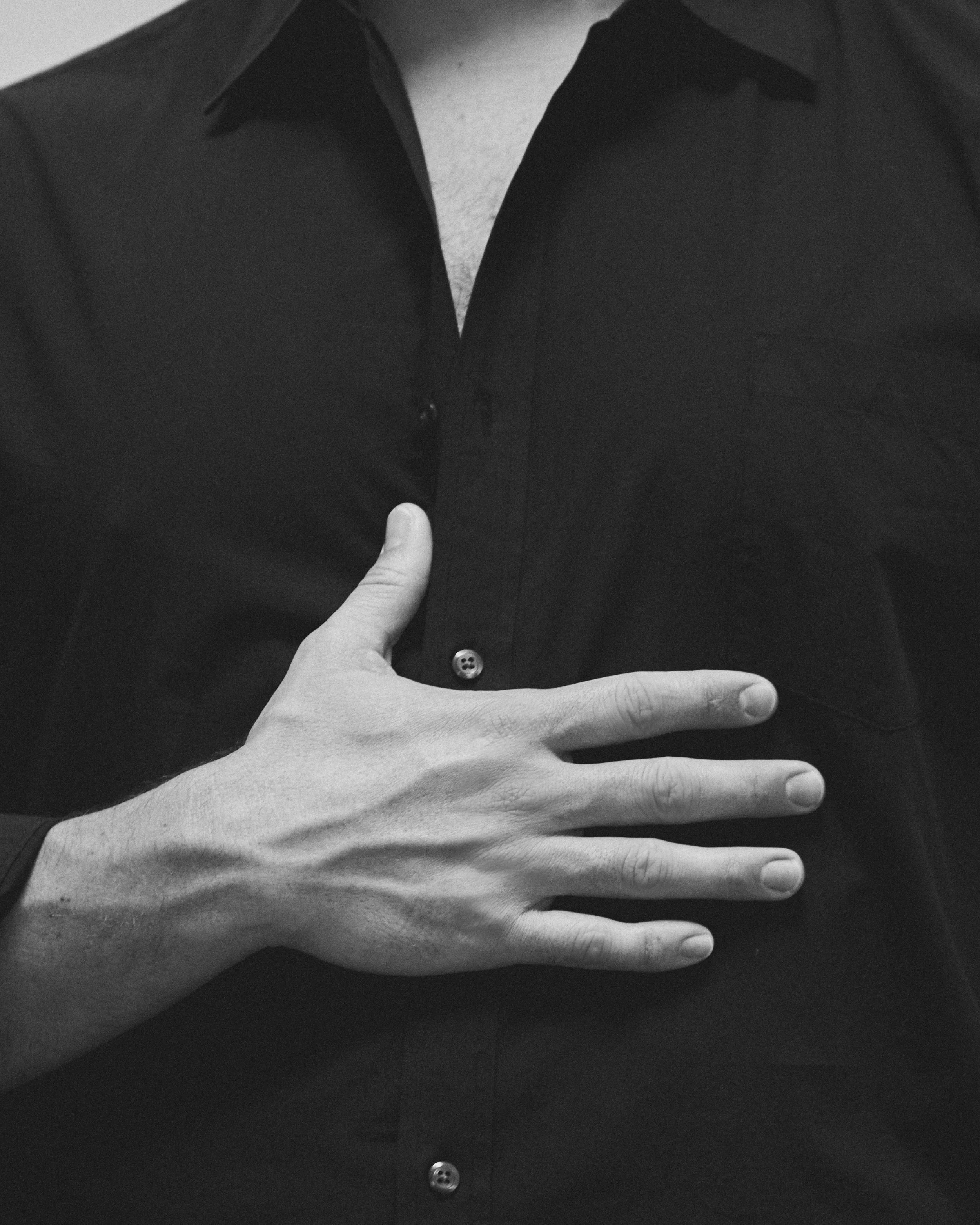 Black and white photo of a person's torso wearing a dark button-up shirt, with one hand placed on their chest, fingers spread out.